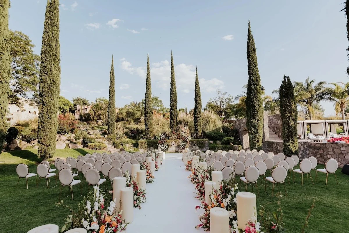 Outdoor wedding venue with white aisle runner, floral arrangements, tall candles, and rows of chairs on a lush green lawn, surrounded by tall cypress trees and landscaping.