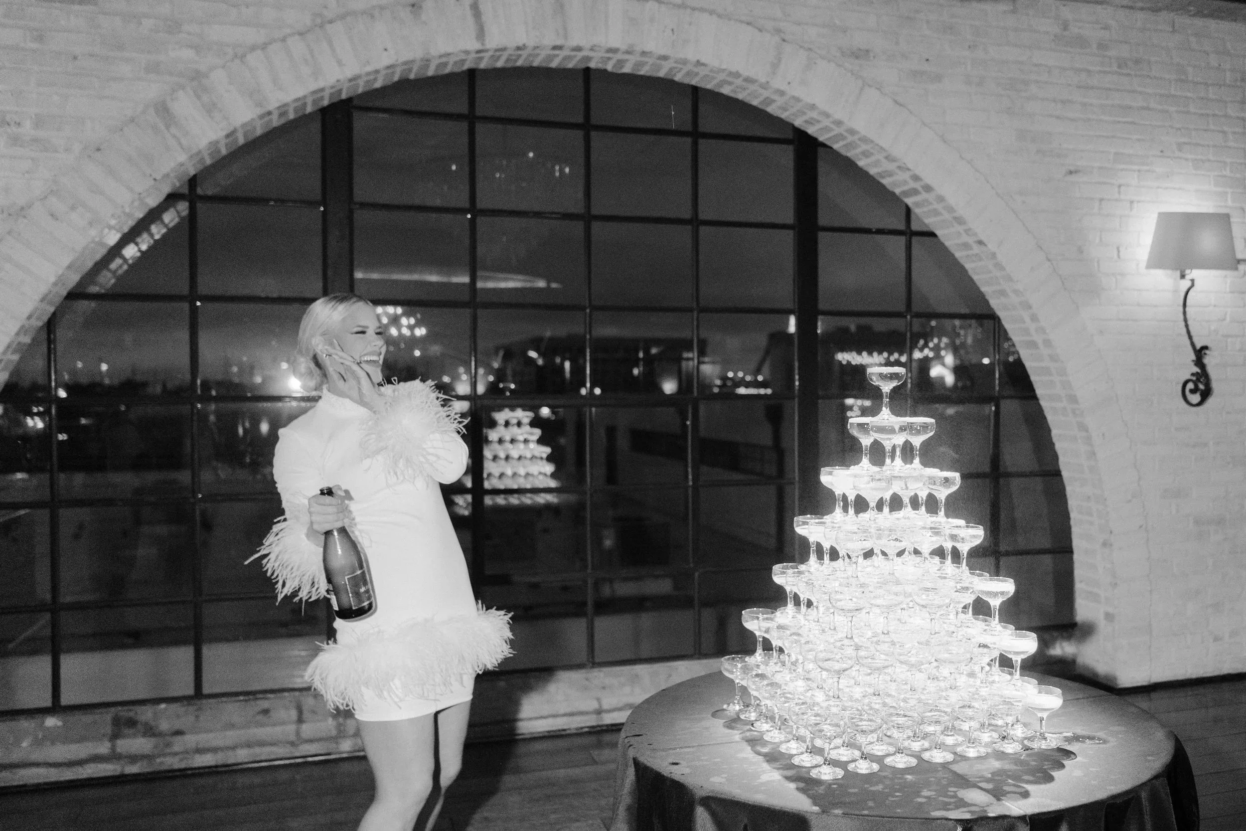 A woman in a feathered dress at an elegant party, smiling and holding a bottle of champagne, standing next to a pyramid of champagne glasses, with a large window and city lights in the background.