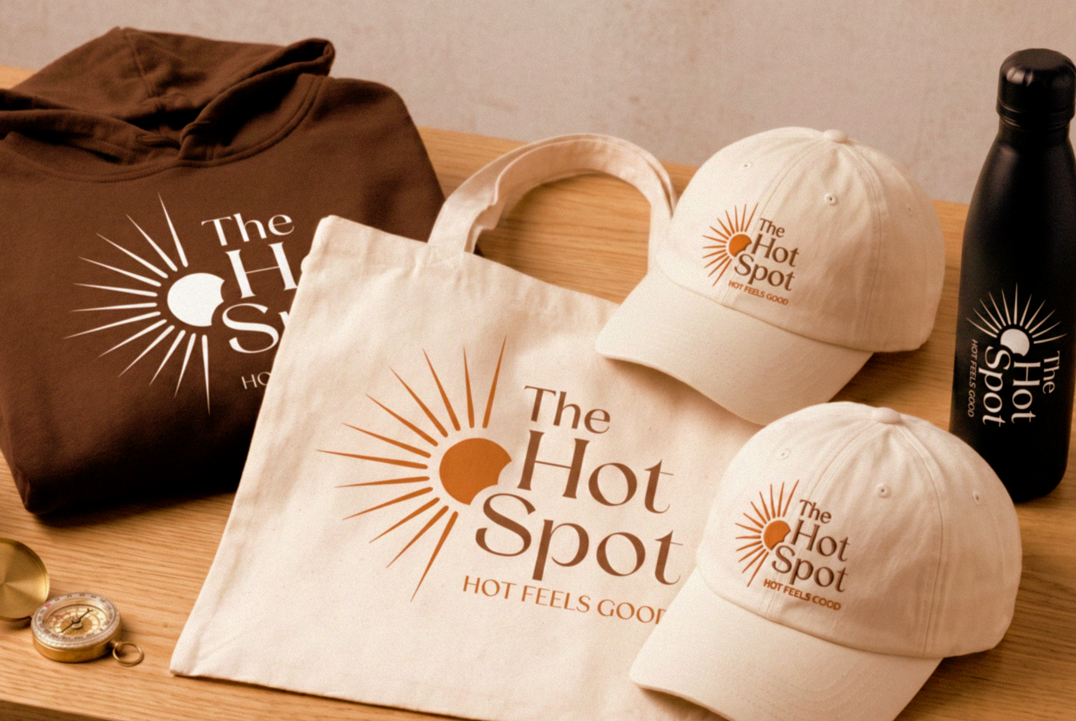 Various merchandise items for The Hot Spot, including a brown hoodie, a white hat, a white canvas tote bag, and a black water bottle, all with the logo and branding of The Hot Spot and the slogan 'Hot Feels Good' on a wooden table.