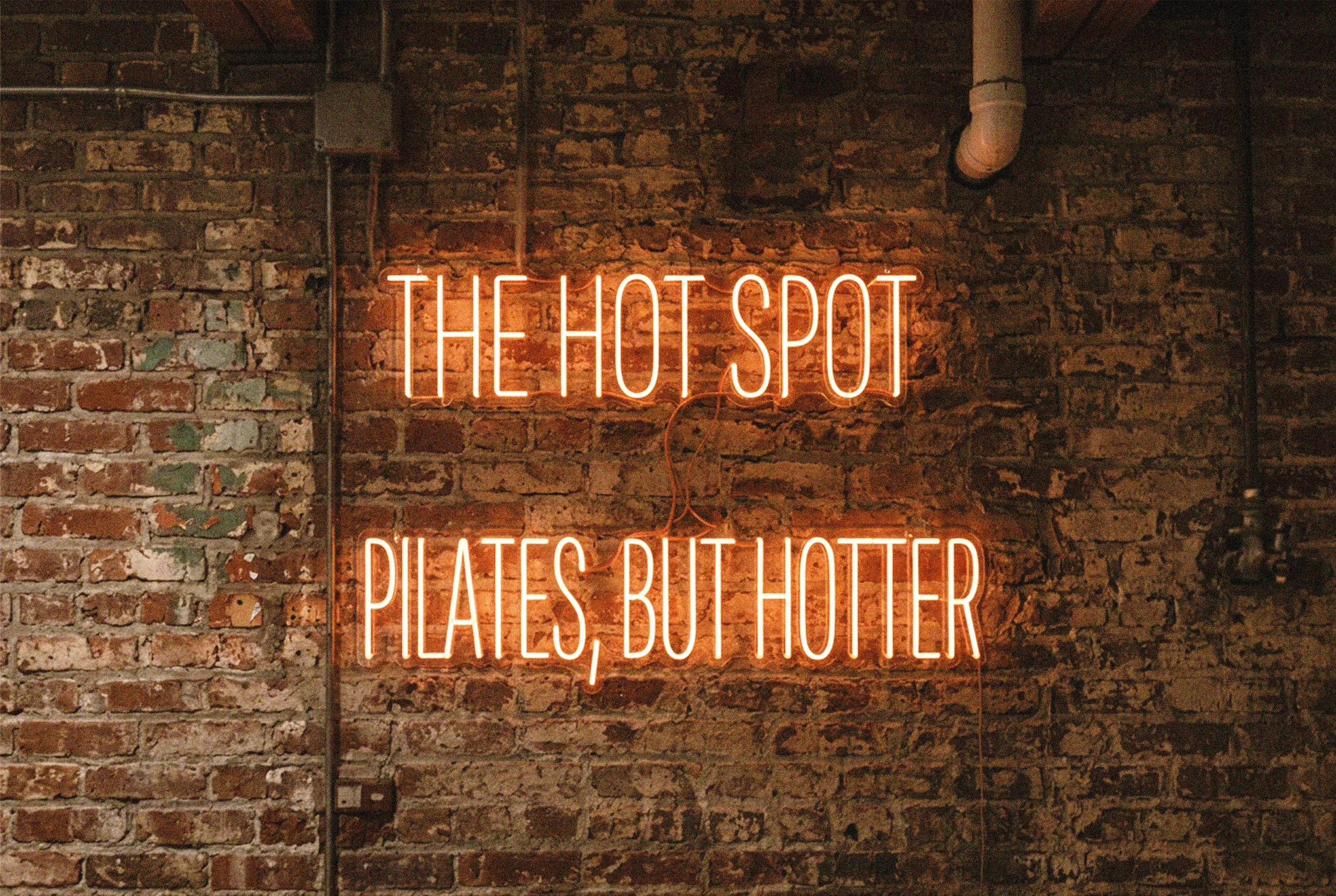 Neon sign reading 'The Hot Spot Pilates, but Hotter' on a brick wall.