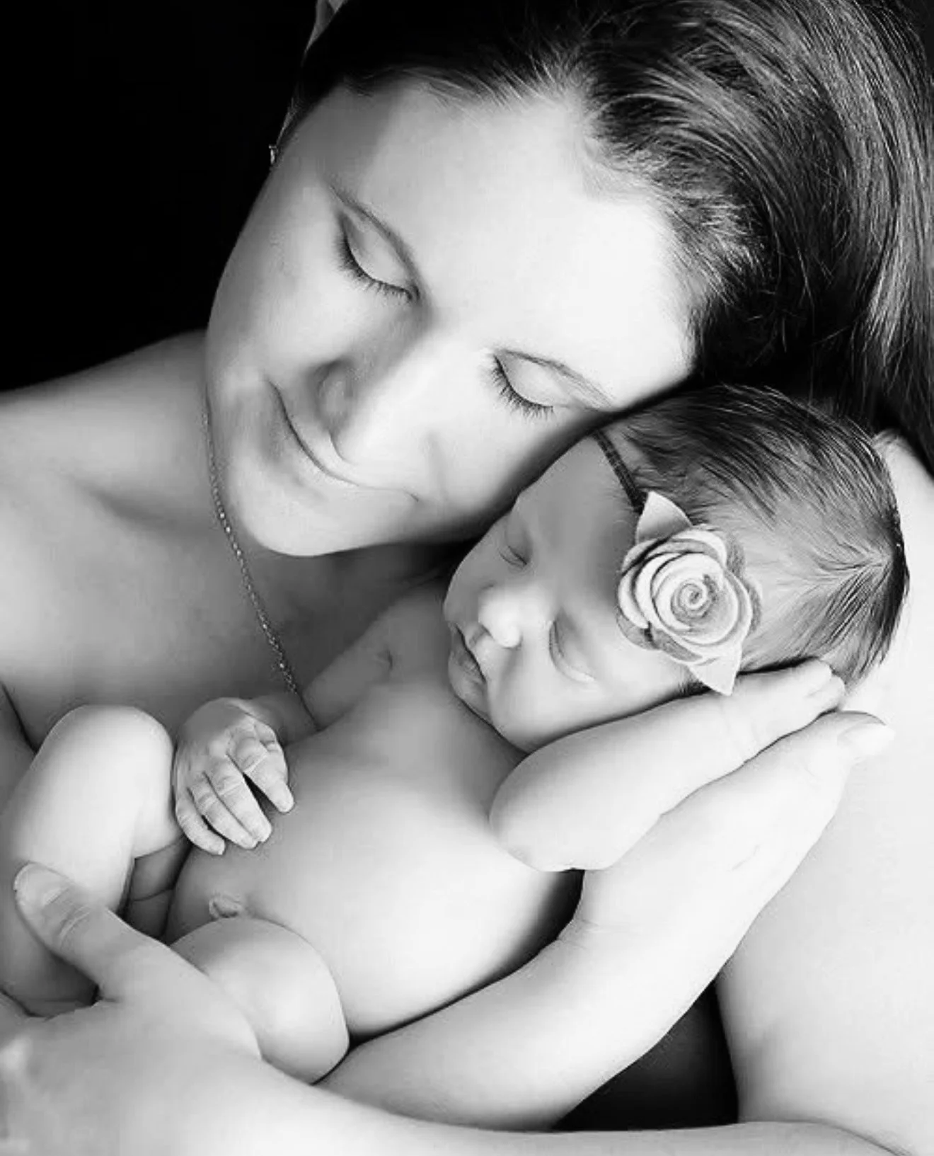 Black and white photo of a woman holding a sleeping baby close to her chest, with both eyes closed. The baby has a flower-shaped headband. The woman has smooth hair and a peaceful expression.