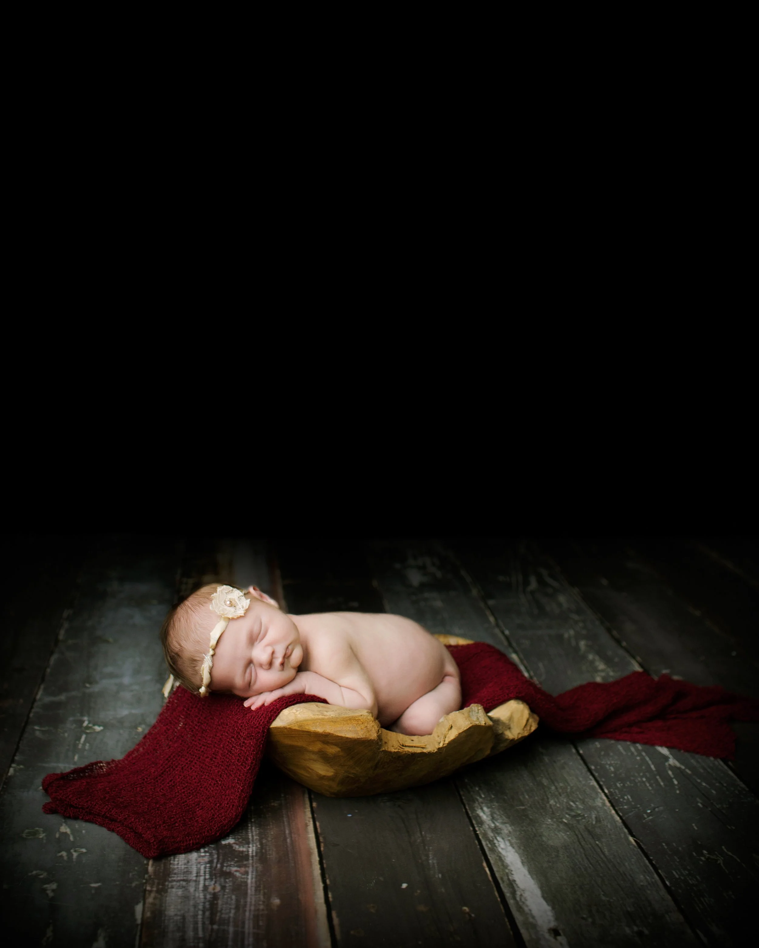 A peacefully sleeping baby lying on a red blanket inside a wooden bowl, with a dark wooden floor beneath and a dark background.