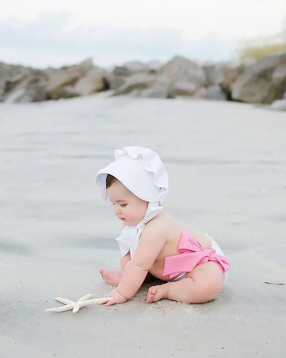 Baby girl sitting on the beach in a white bonnet and pink swimsuit with a large pink bow, playing with a starfish.