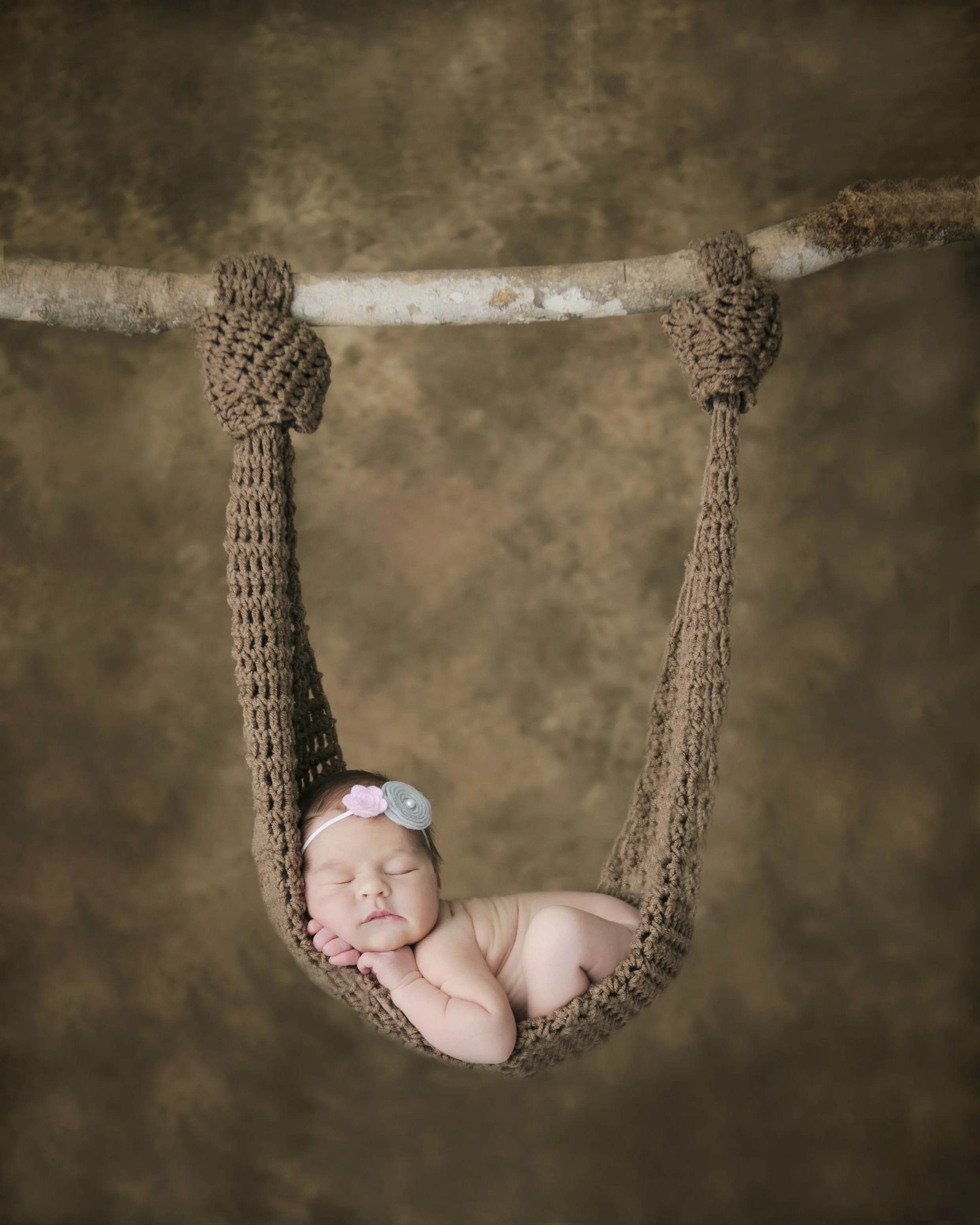 A sleeping baby girl wearing a headband with flowers, curled up in a woven hammock with a wooden support, against a brown textured background.