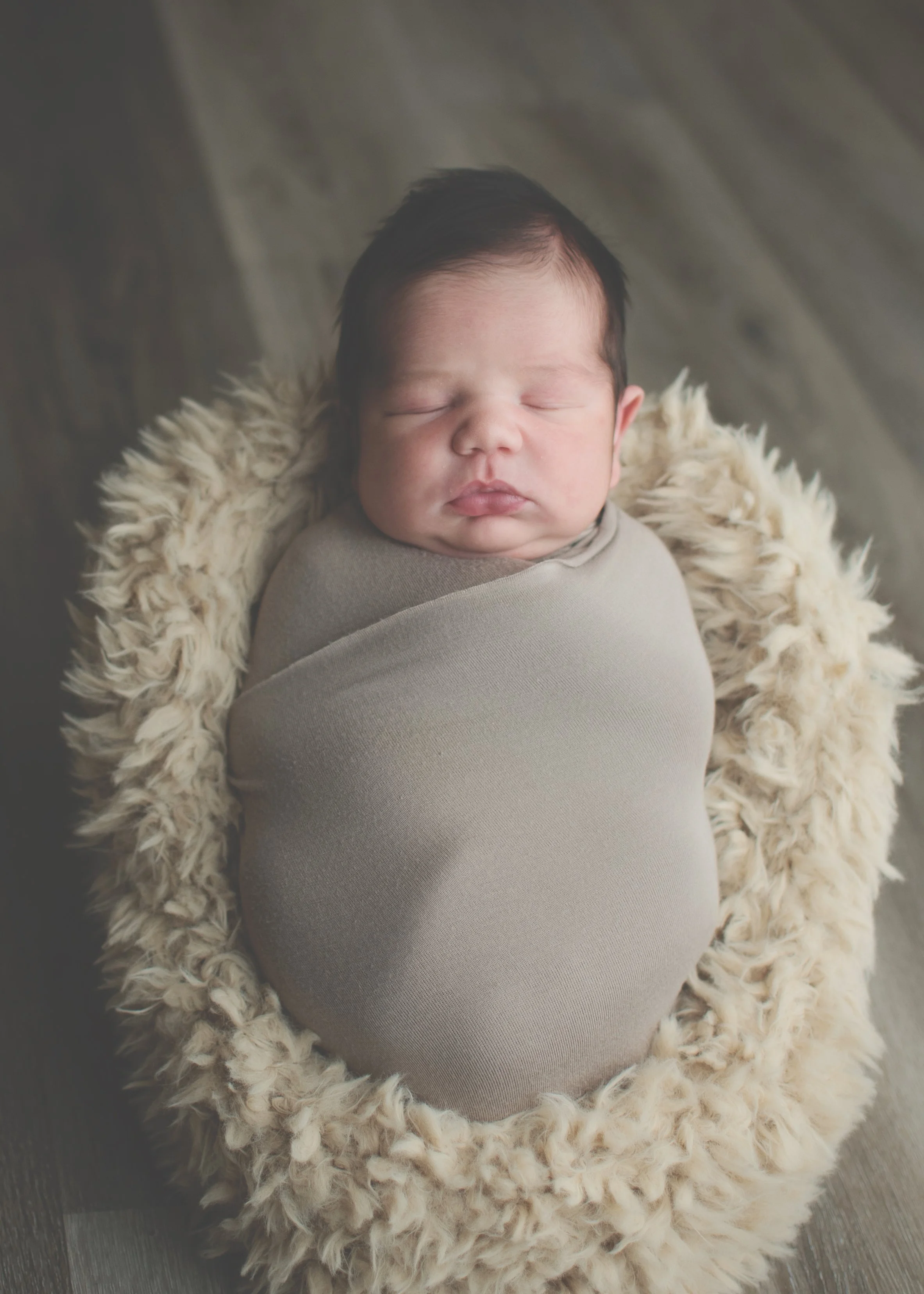 A newborn baby with closed eyes, wrapped in a beige blanket, lying on a fluffy cream-colored rug on a wooden floor.