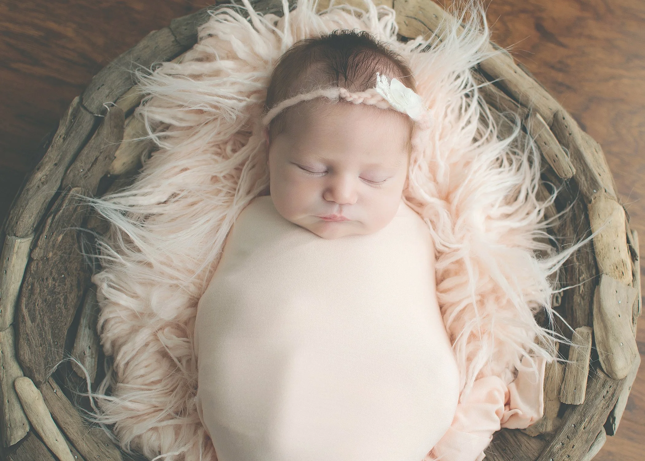 Close-up of a sleeping newborn baby with dark hair, wearing a beige outfit, lying on a soft pink fluffy blanket inside a woven wooden basket on a wooden surface.