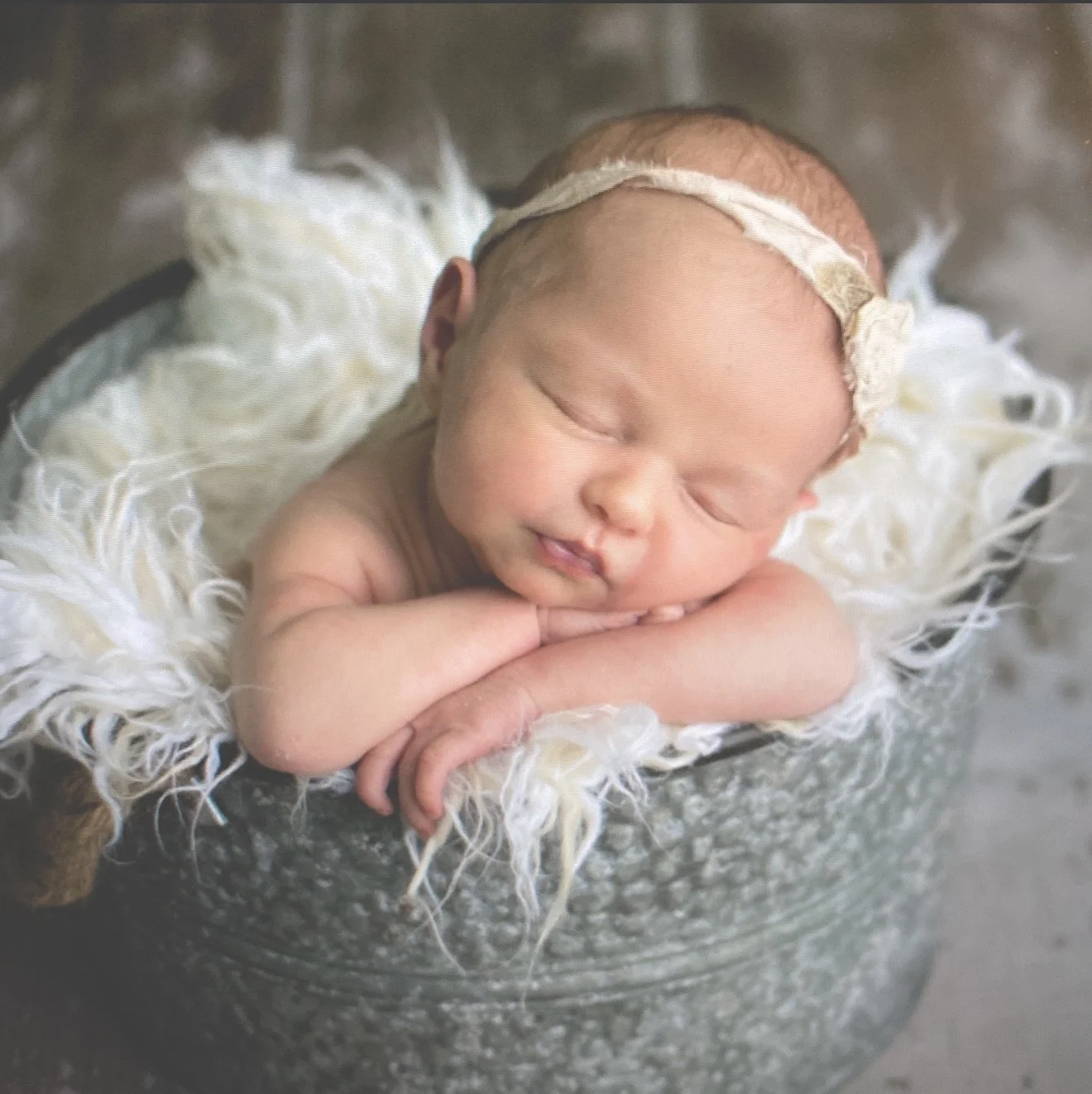 A sleeping newborn baby with a headband, resting on a fluffy white blanket in a metal bowl.