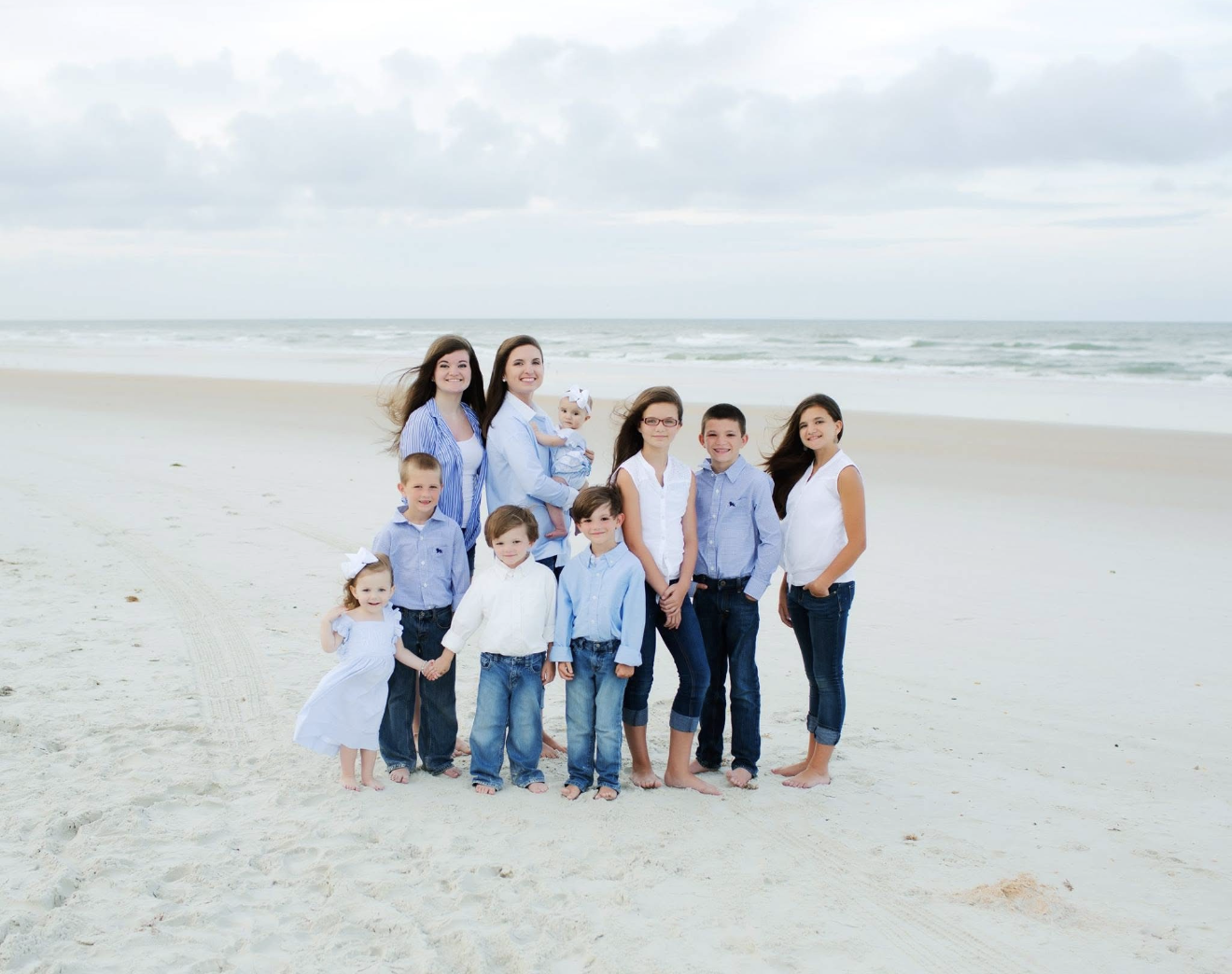 Family of ten, including children and adults, dressed in casual blue and white clothing, posing on the beach with sand and ocean waves in the background.