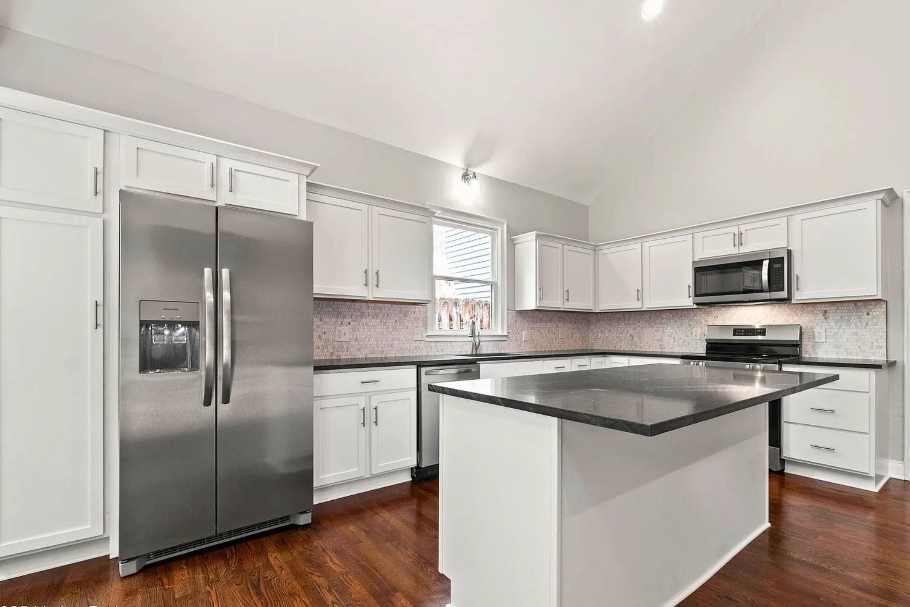 Modern kitchen with white cabinets, stainless steel refrigerator, black countertops, pink tile backsplash, and hardwood floors.