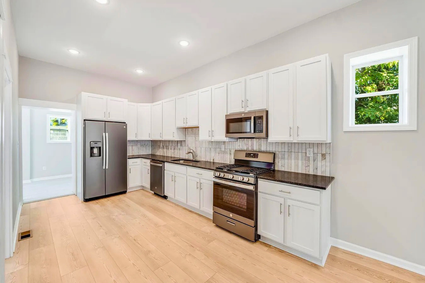 A modern kitchen with white cabinets, stainless steel appliances including a refrigerator, microwave, and oven, light wood flooring, a gray tiled backsplash, a sink under a window, and recessed ceiling lights.