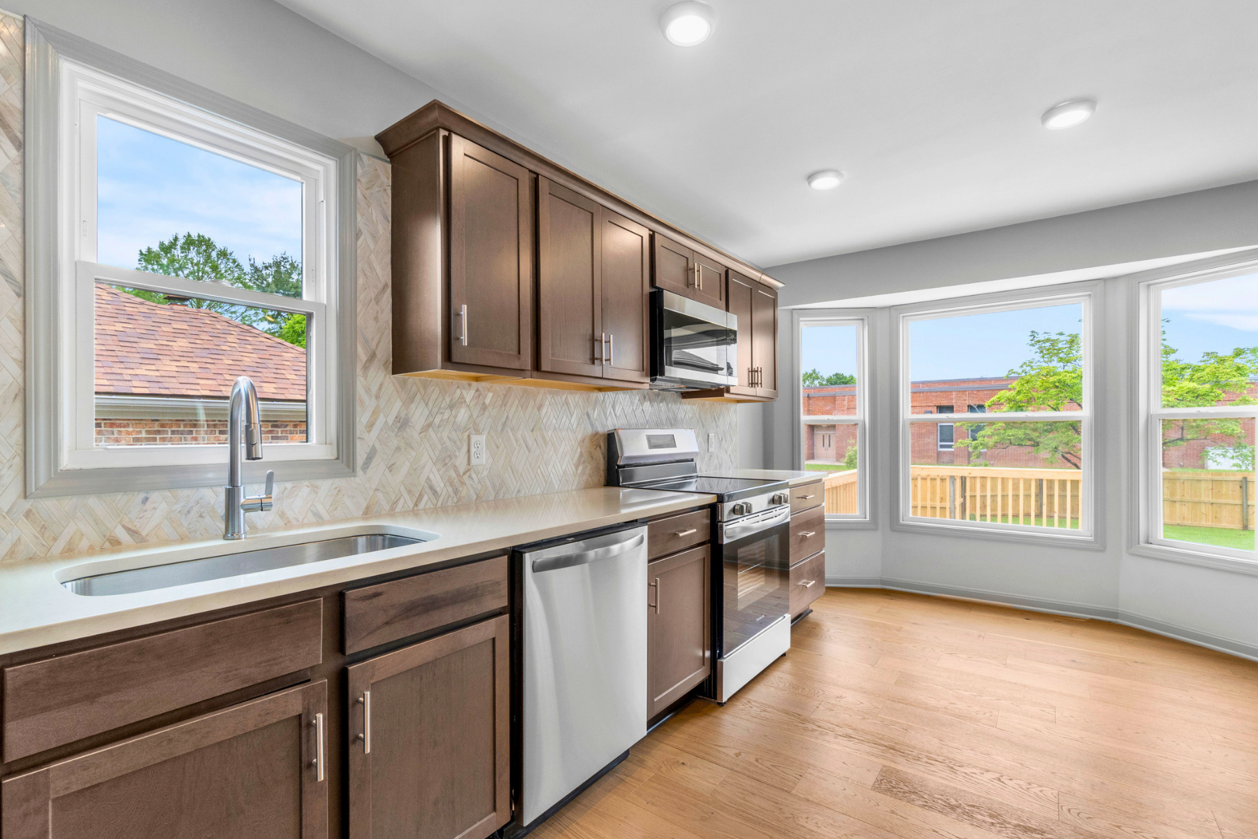 Modern kitchen with dark brown cabinets, a beige countertop, a stainless steel sink under a window, a stainless steel microwave above a stove, a dishwasher, and large windows offering a view of a backyard with a wooden fence and trees.