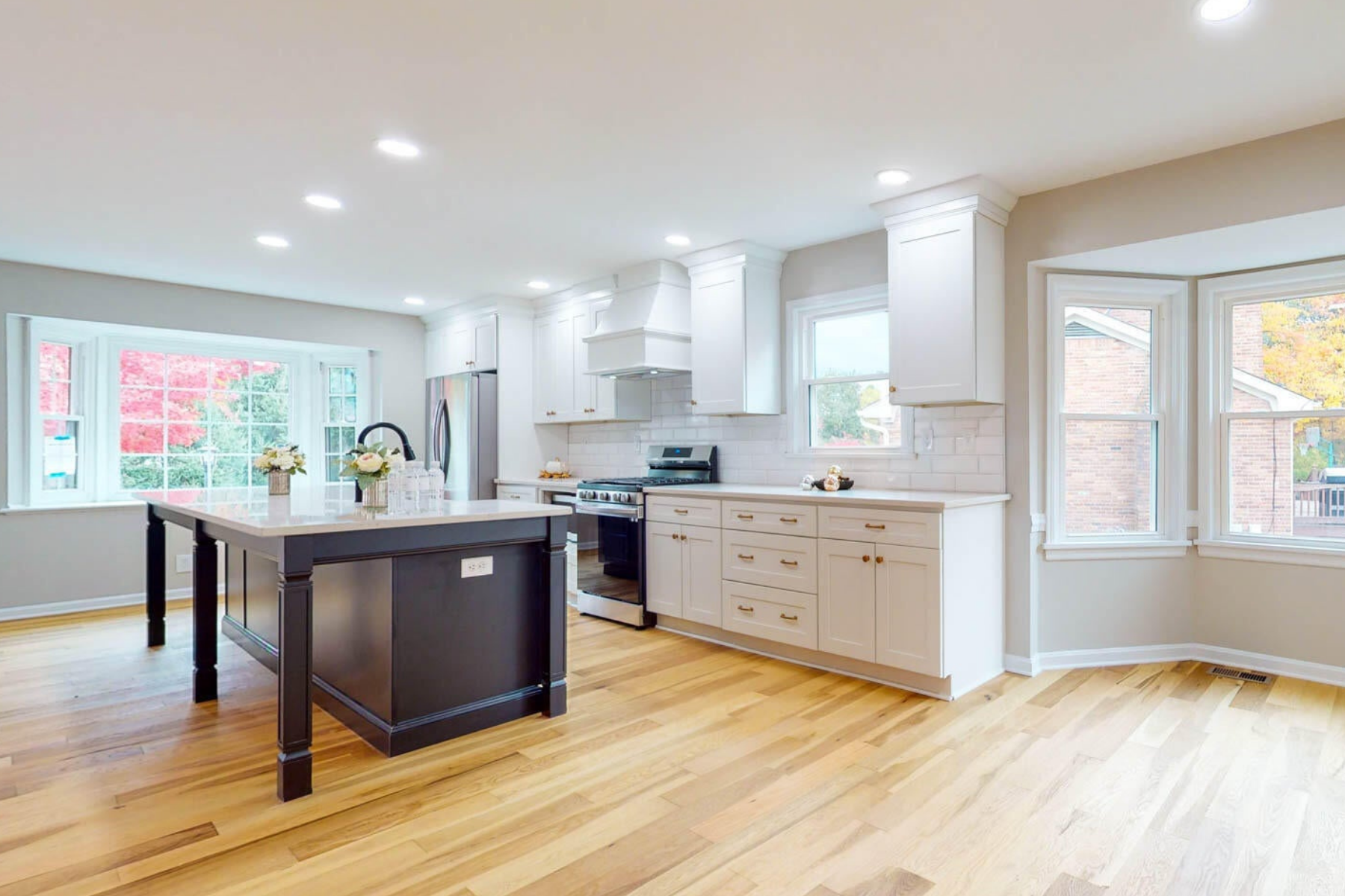 Modern, bright kitchen featuring white cabinets, stainless steel appliances, a black kitchen island, large windows with a view of trees and a brick house, and hardwood flooring.