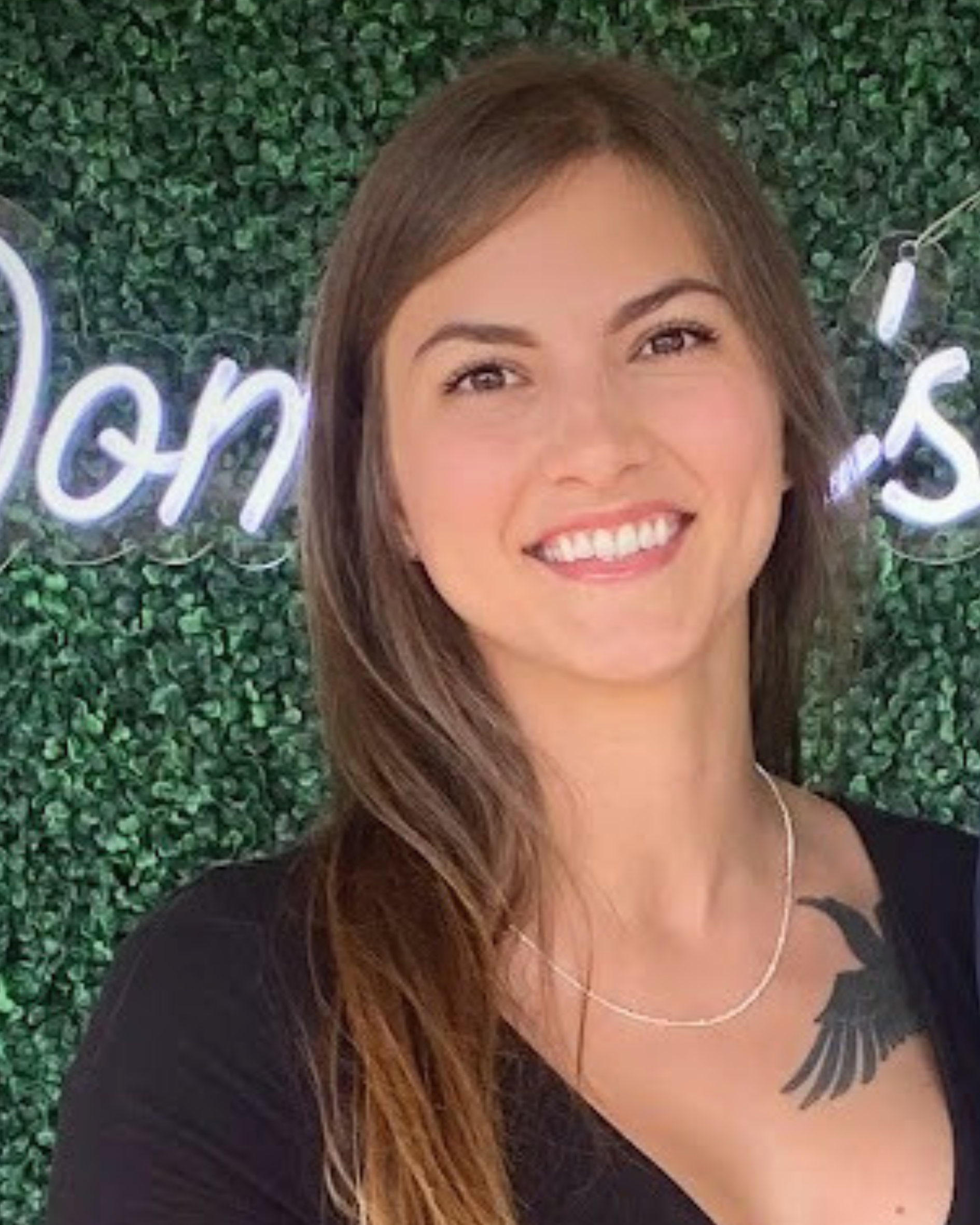 Young woman with brown hair, smiling, wearing a black top, standing in front of a green leafy background with a neon sign that has partially visible words.
