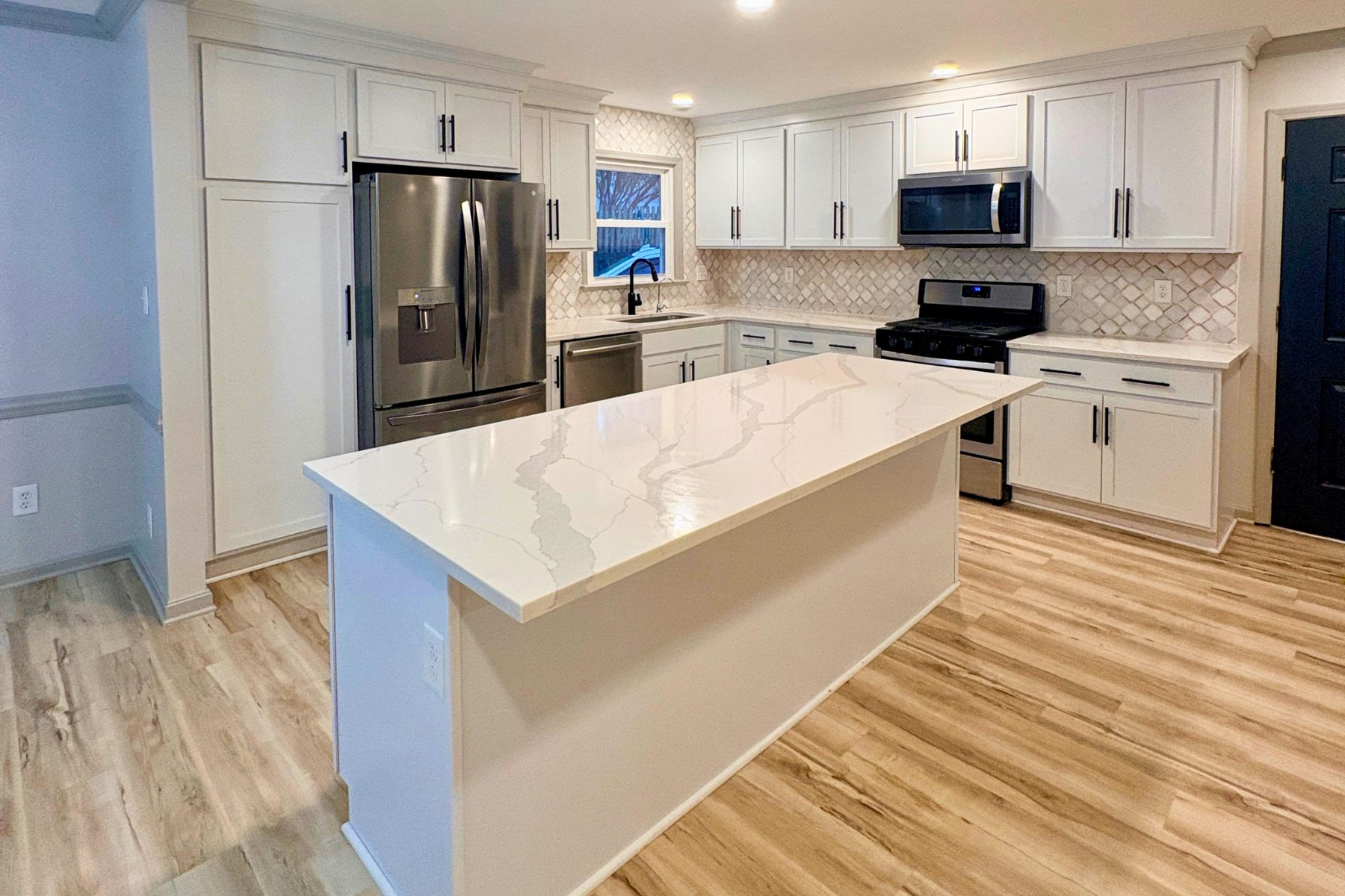 Modern kitchen with white cabinets, stainless steel appliances including a refrigerator, microwave, oven, and dishwasher. Light-colored stone countertop island and hardwood floors. Window above sink showing outdoor view.