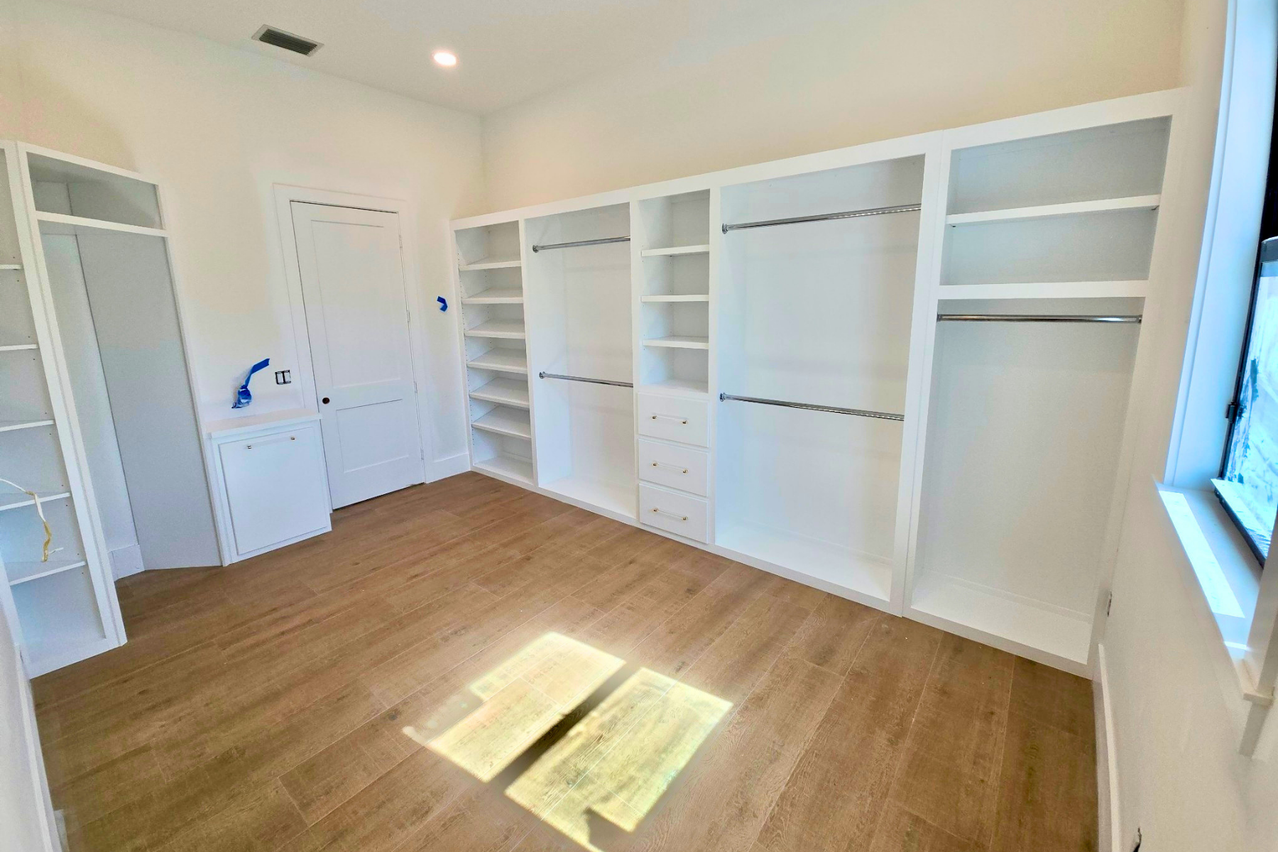 Empty walk-in closet with built-in white shelving and hanging rods, a window on the right, and a wood floor.