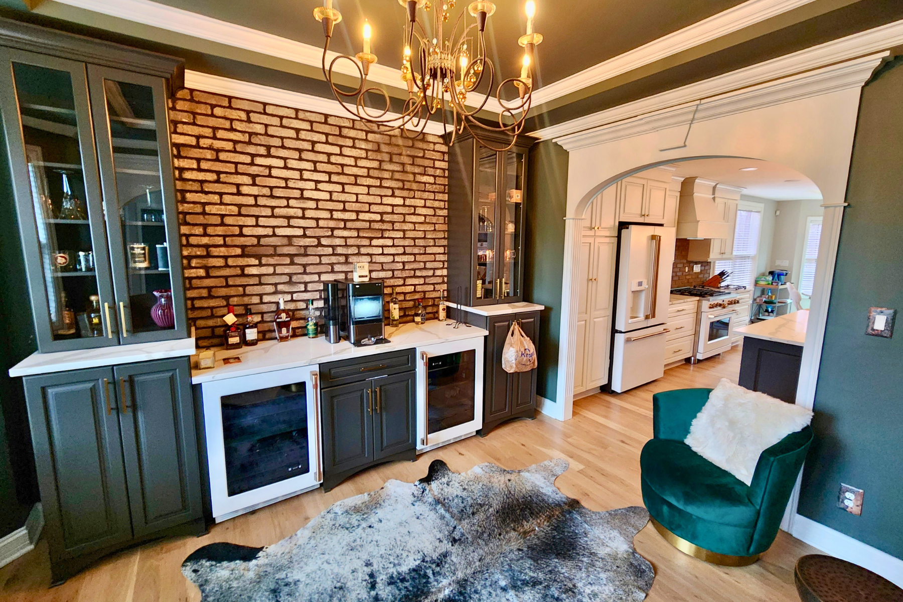 A kitchen with an exposed brick wall, dark and white cabinetry, a chandelier, a bar area with bottles, and an adjoining space with a white stove, refrigerator, and large windows. There's a green velvet armchair with a white fur throw, a cowhide rug, 