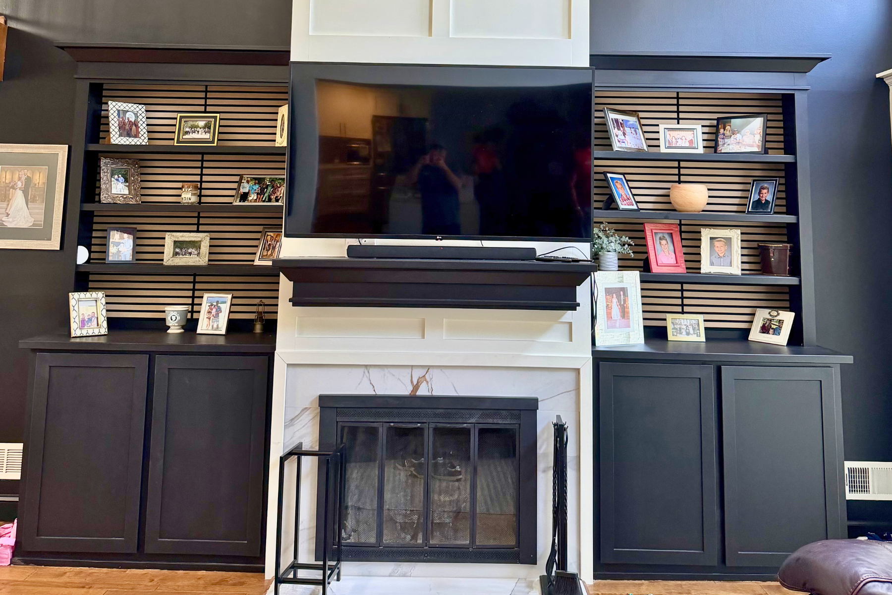 Living room fireplace with black cabinets on either side, a black mantel, a large flat-screen TV above the mantel, and a collection of family photos on black and white shelves surrounding the TV.