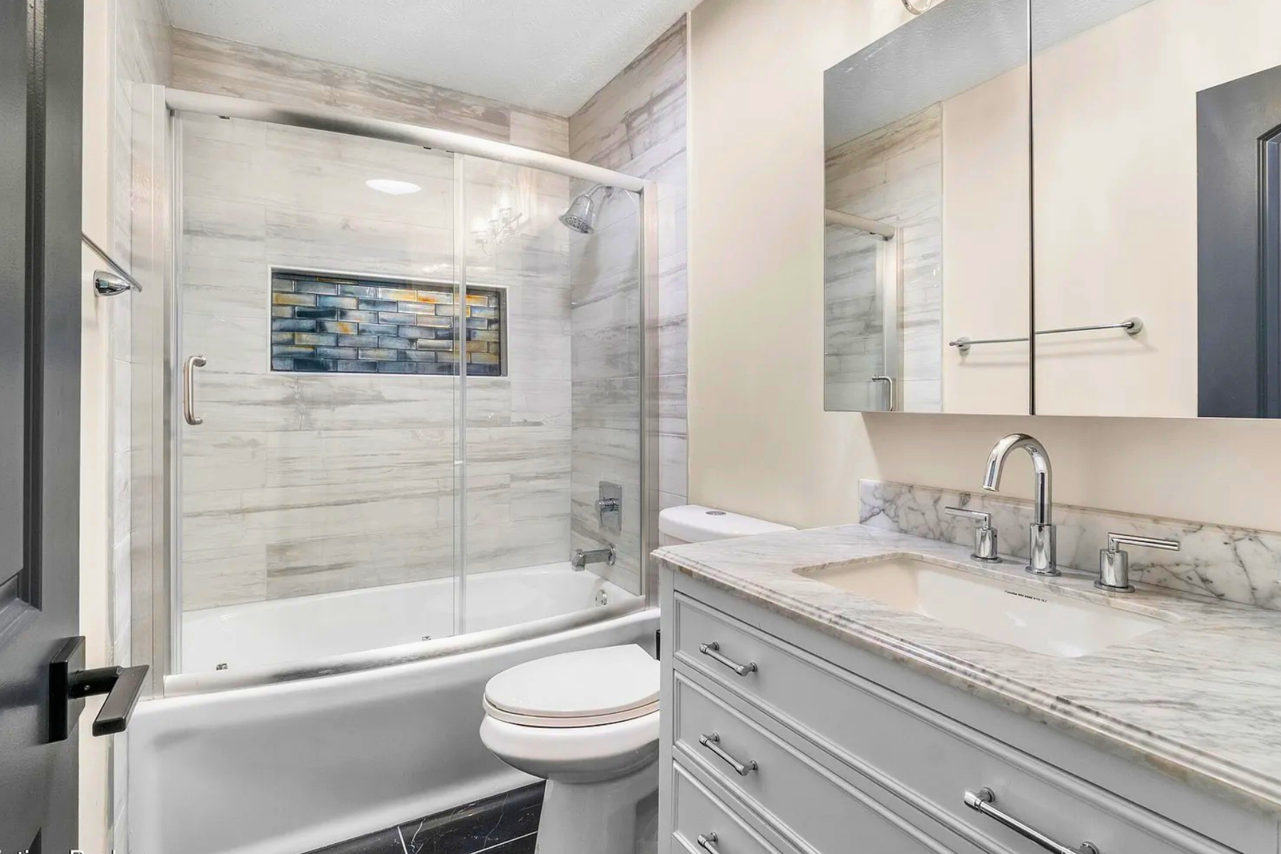 A modern bathroom featuring a glass-enclosed shower with a built-in tiled wall niche, marble countertops, a mirror cabinet, a white toilet, and a marble vanity with a silver faucet, all with neutral-colored walls and flooring.