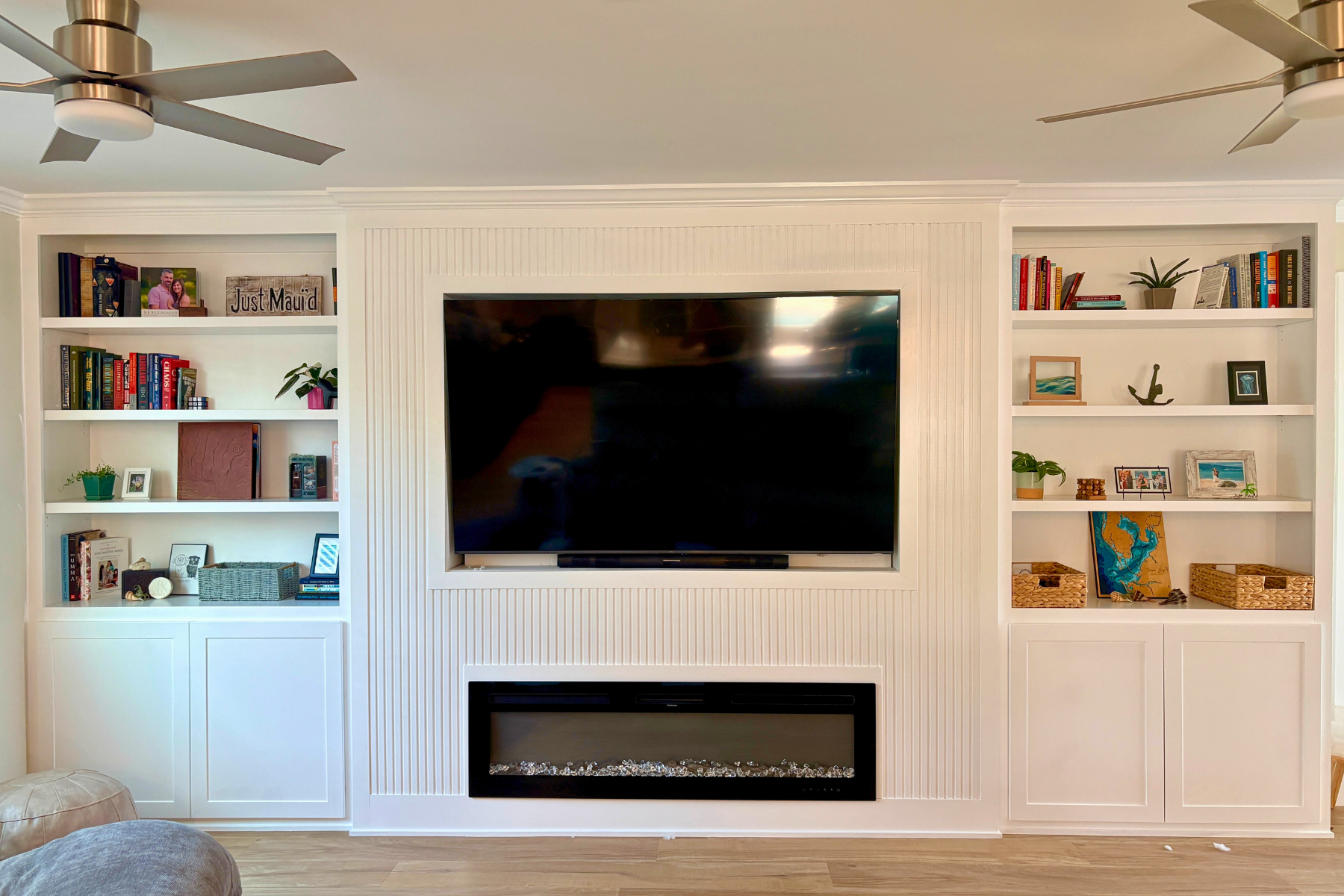 Living room with white built-in shelves on either side of a wall-mounted flat-screen TV and a rectangular electric fireplace below, with various books, plants, and decorative items on the shelves.