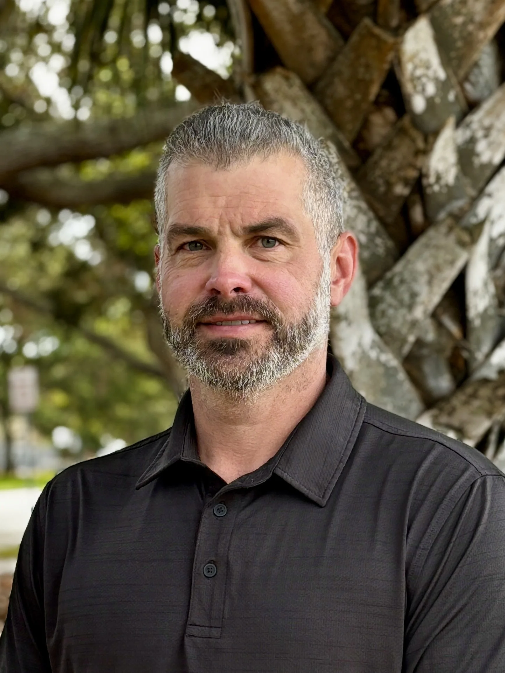 Close-up of a middle-aged man with salt-and-pepper hair and beard, wearing a black shirt, standing outdoors near a tree with green leaves and blurry background.