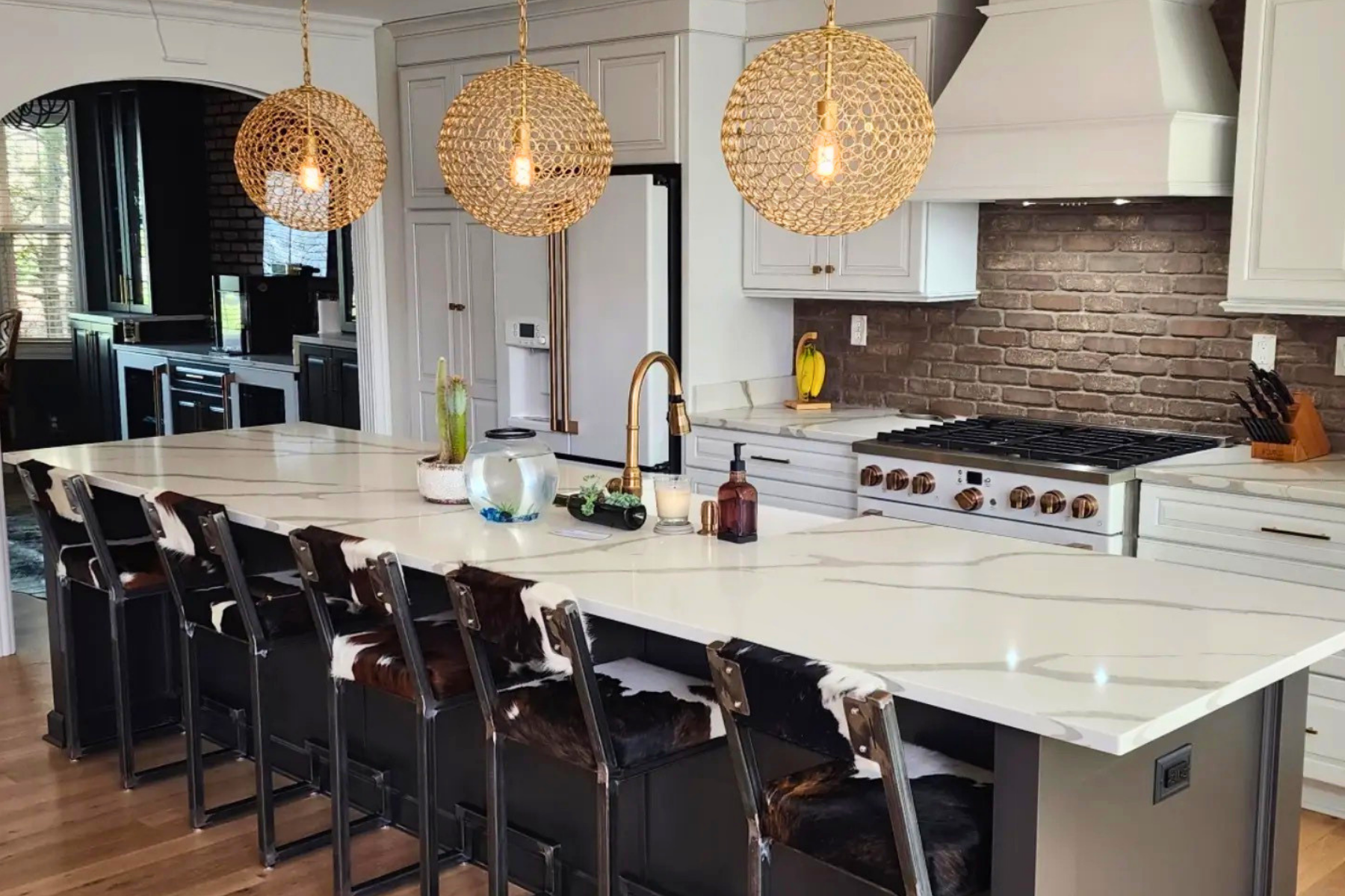 Modern kitchen with white cabinetry, brick backsplash, and a large white island. Three woven pendant lights hang above the island. The kitchen has a gold faucet, decorative items, and a row of black and white cowhide bar stools.