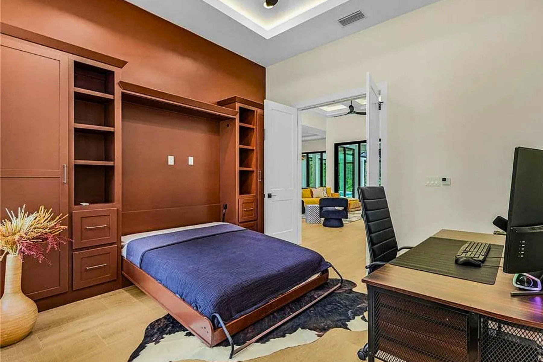 A bedroom with a wooden built-in wall unit, a bed with a purple cover, a vase with flowers, a desk with a computer and chair, and a view into a living room with yellow and gray furniture and large windows.