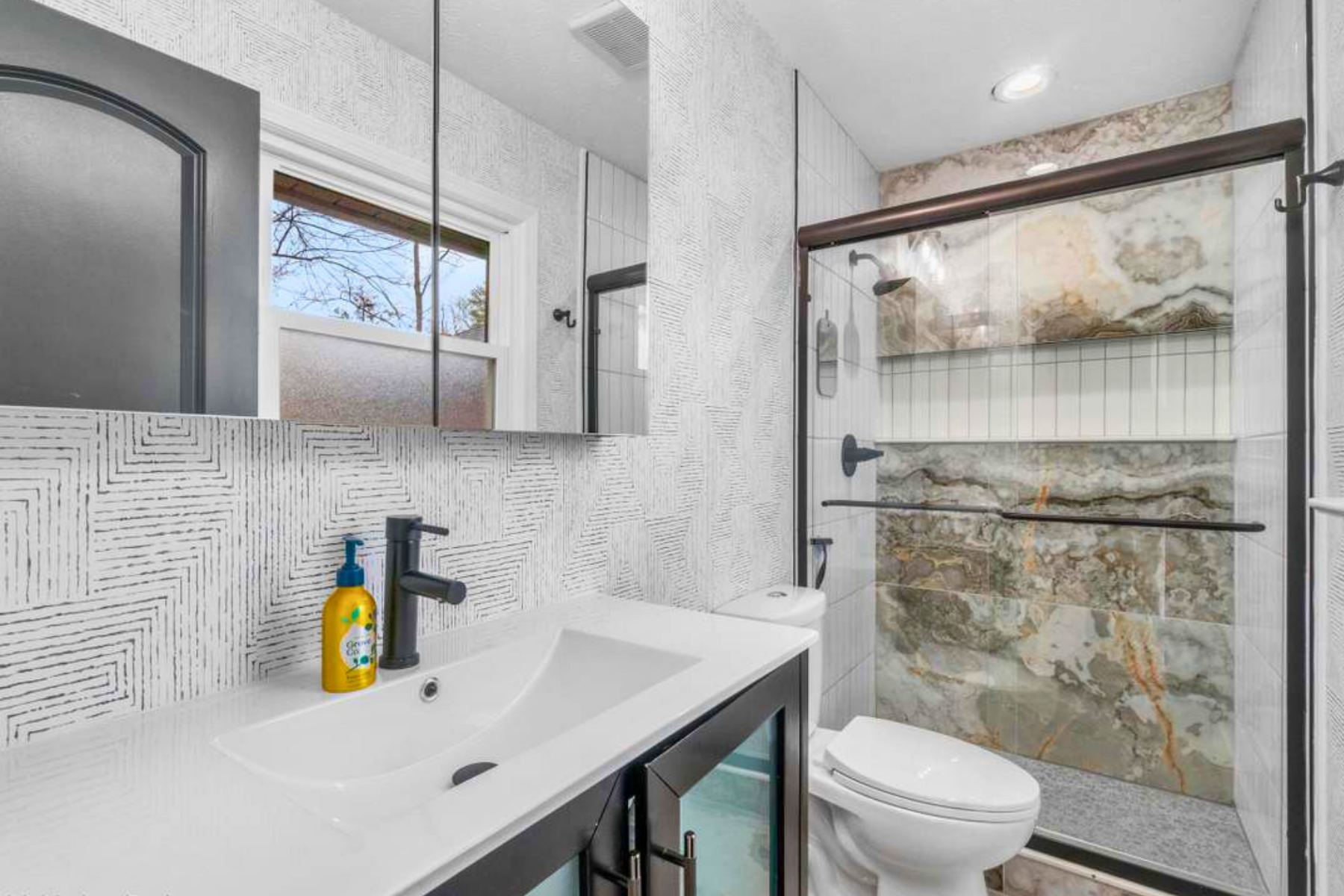 Modern bathroom with a white sink, black faucet, and a yellow soap dispenser. The wall above the sink has textured white tile, and a mirror reflects a window with a view of trees outside. A toilet is positioned next to a shower area with marble tiles