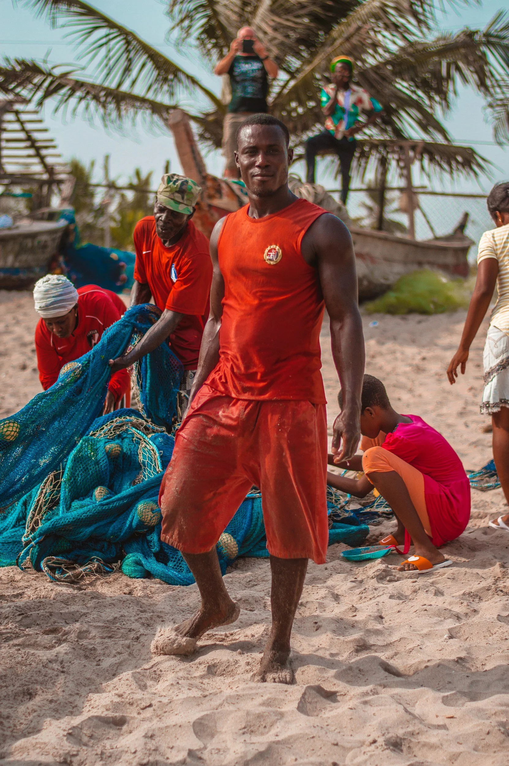 Young man walking on the beach with sand in the foreground, surrounded by people sitting and working with fishing nets, palm trees in the background, and a boat.