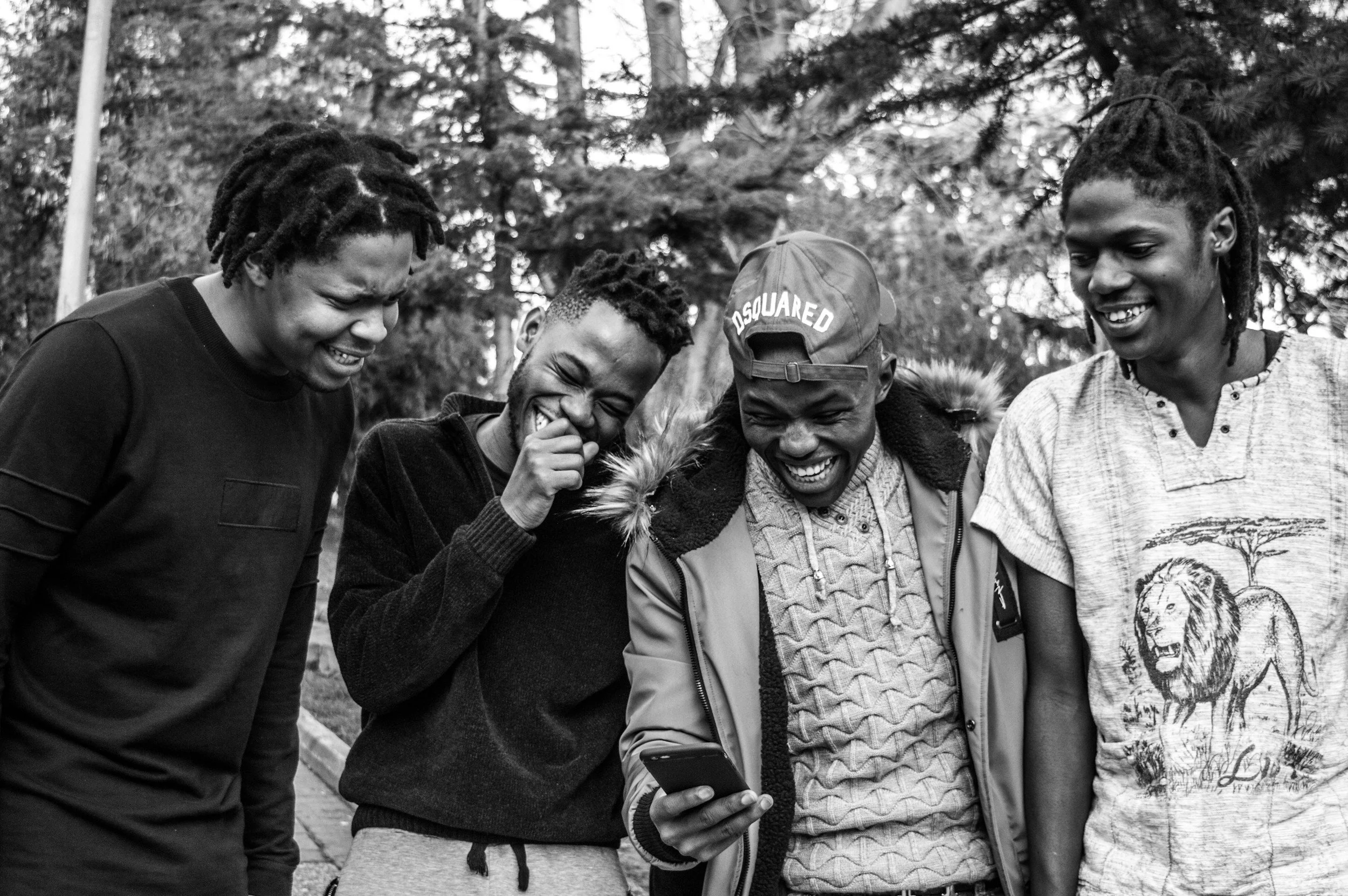 Five young men with dreadlocks smiling and looking at a phone, standing outdoors among trees.