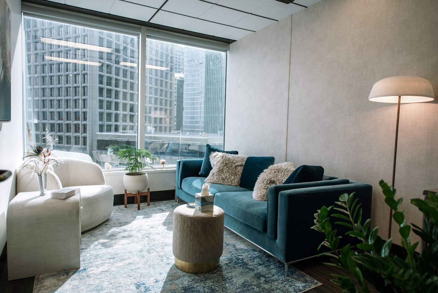 Modern living room with large window showing city buildings, a blue sofa with fluffy pillows, a white armchair, a small round beige ottoman, a potted plant, a tall floor lamp, and a patterned rug.
