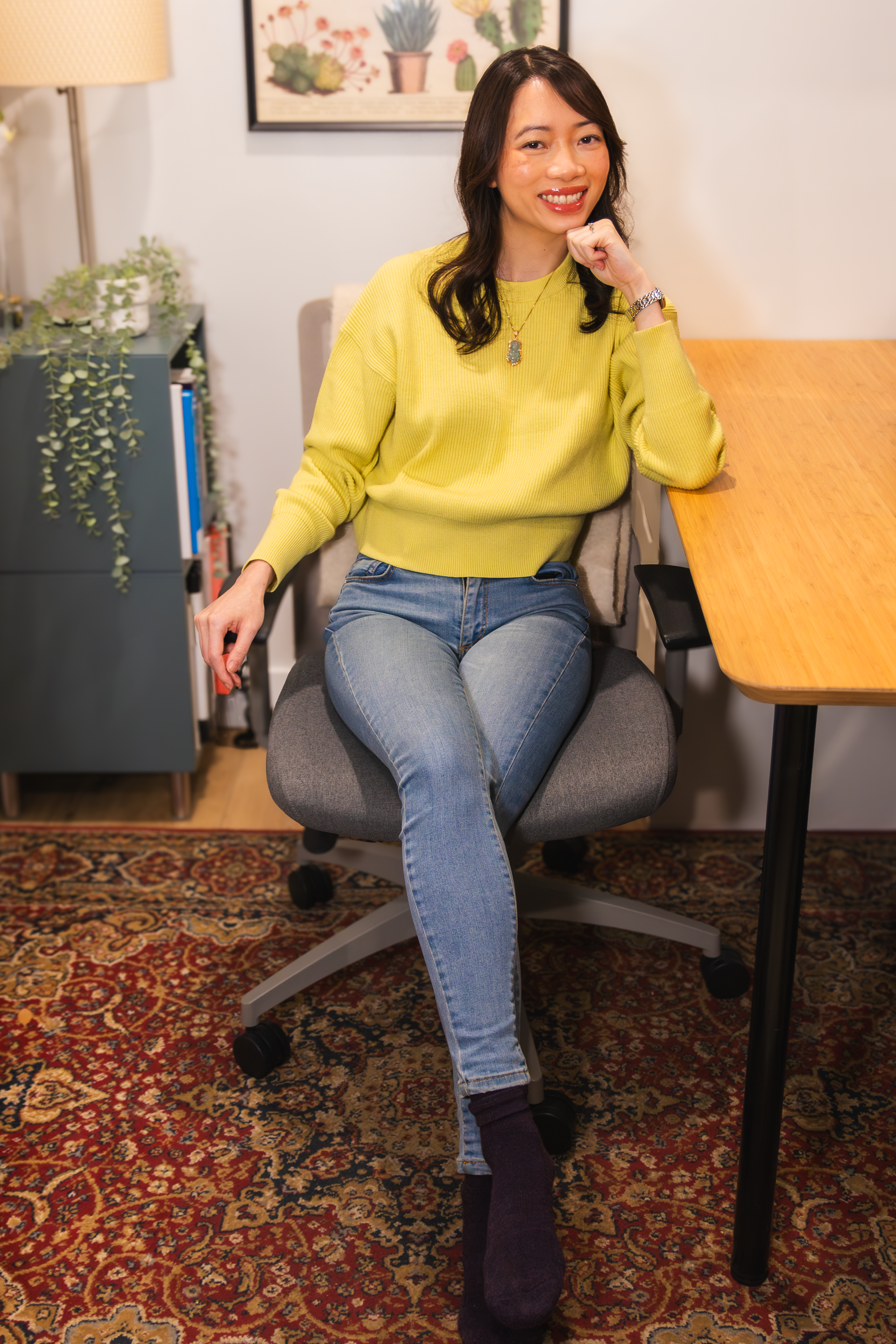 A woman with long dark hair wearing a yellow sweater sitting on a gray office chair in a room with a wooden table, patterned rug, framed artwork of cacti, a green storage cabinet with plants, and a tall lamp.