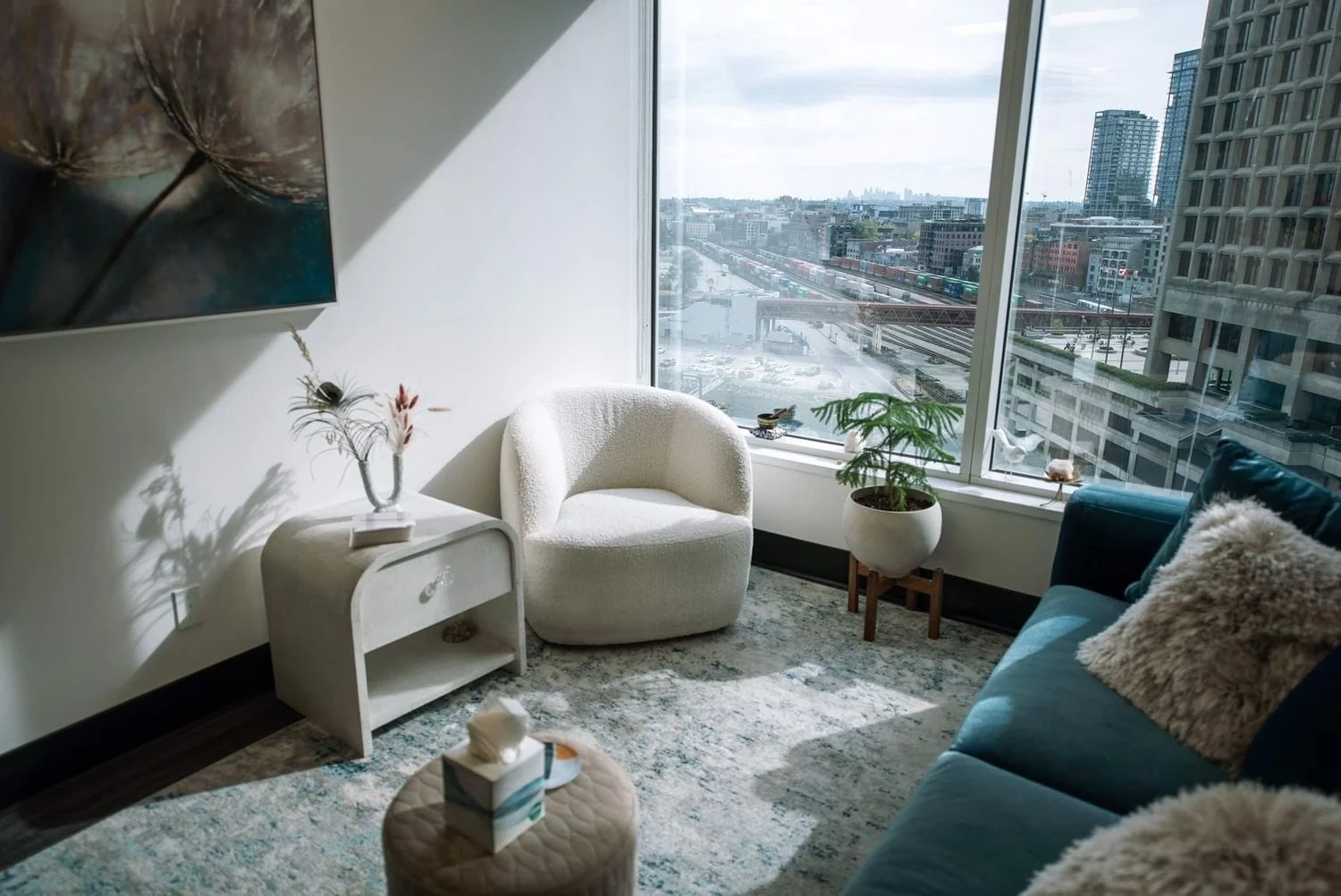 Living room with large window showing cityscape, white armchair, blue sofa with pillows, potted plant, side table with decorative items, and wall art