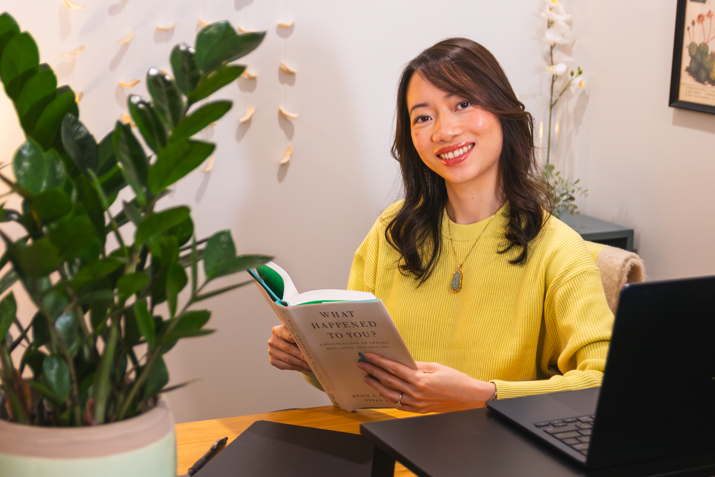 A woman with shoulder-length brown hair, wearing a yellow sweater, smiling while reading a book titled 'What Happened to You?' in an indoor setting with plants and framed art on the wall.