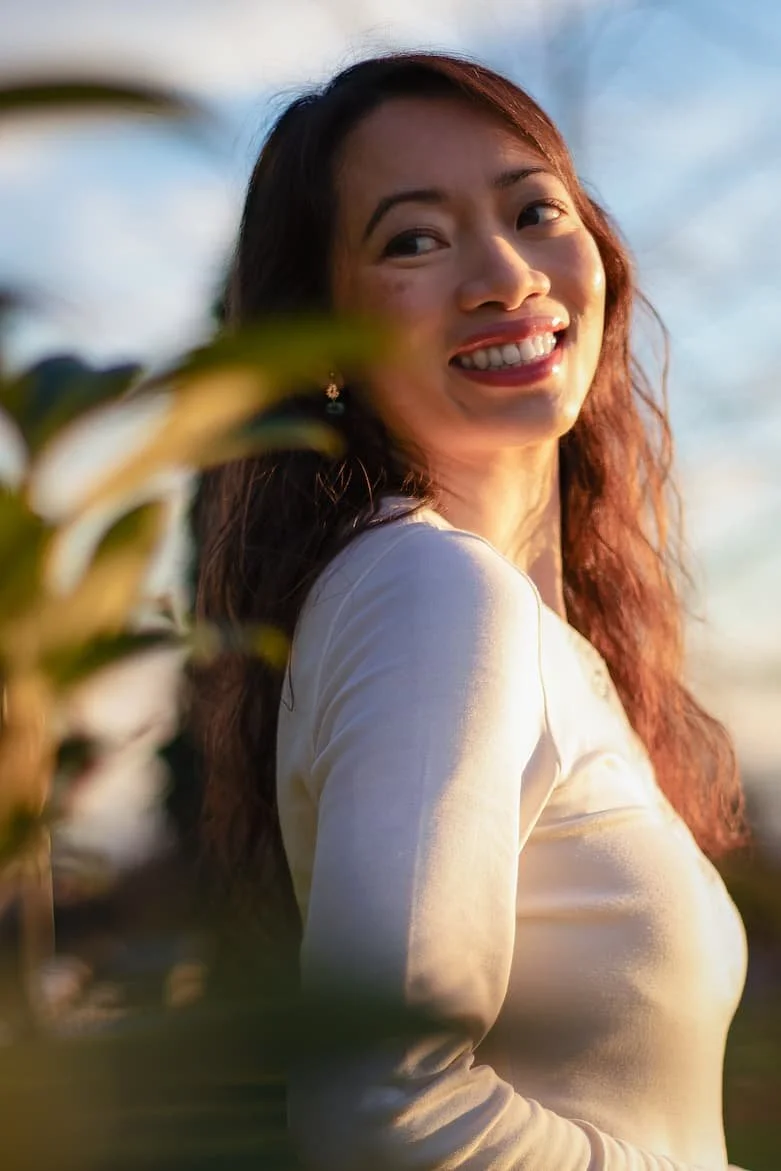 A woman with long, wavy brown hair smiling outdoors during golden hour, wearing a light-colored long sleeve top, with blurred green leaves and a sky in the background.
