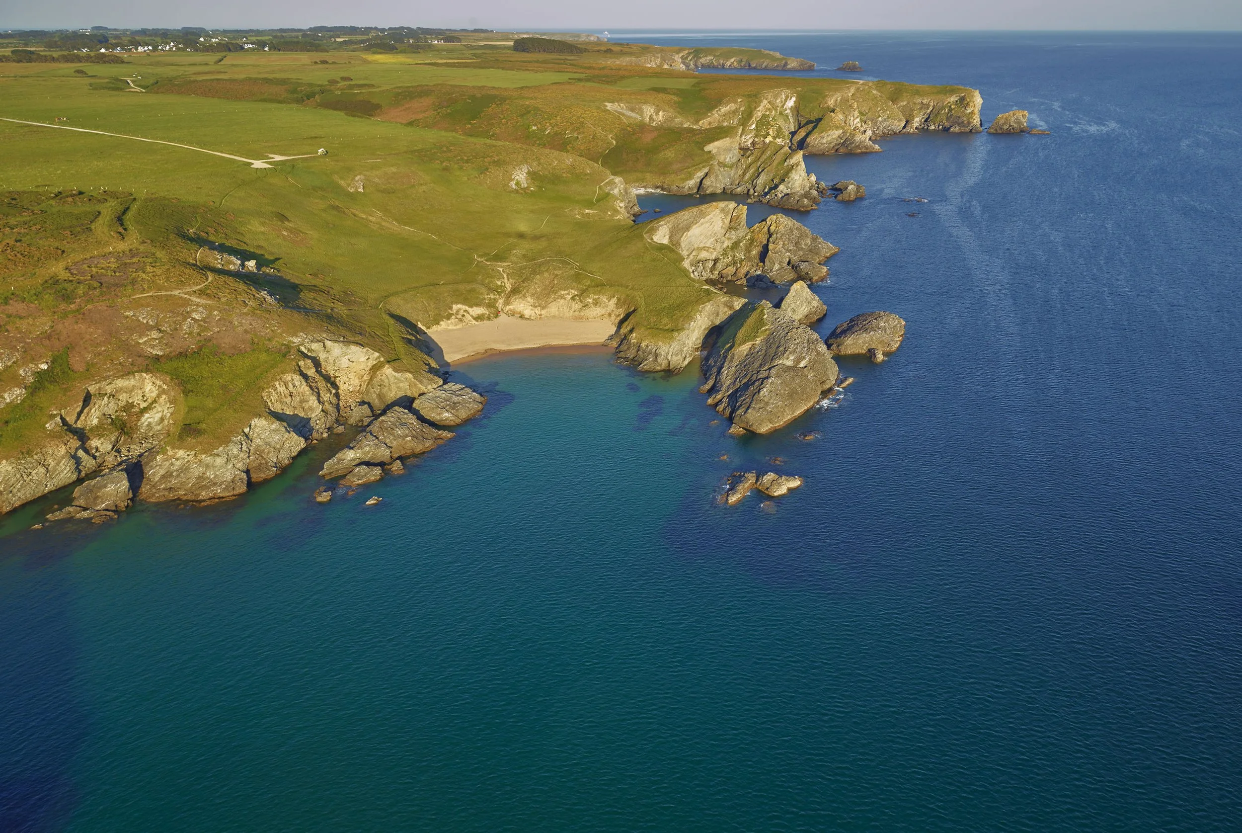 Paysage côtier avec des falaises rocheuses, une plage, de la mer et du terrain verdoyant.
