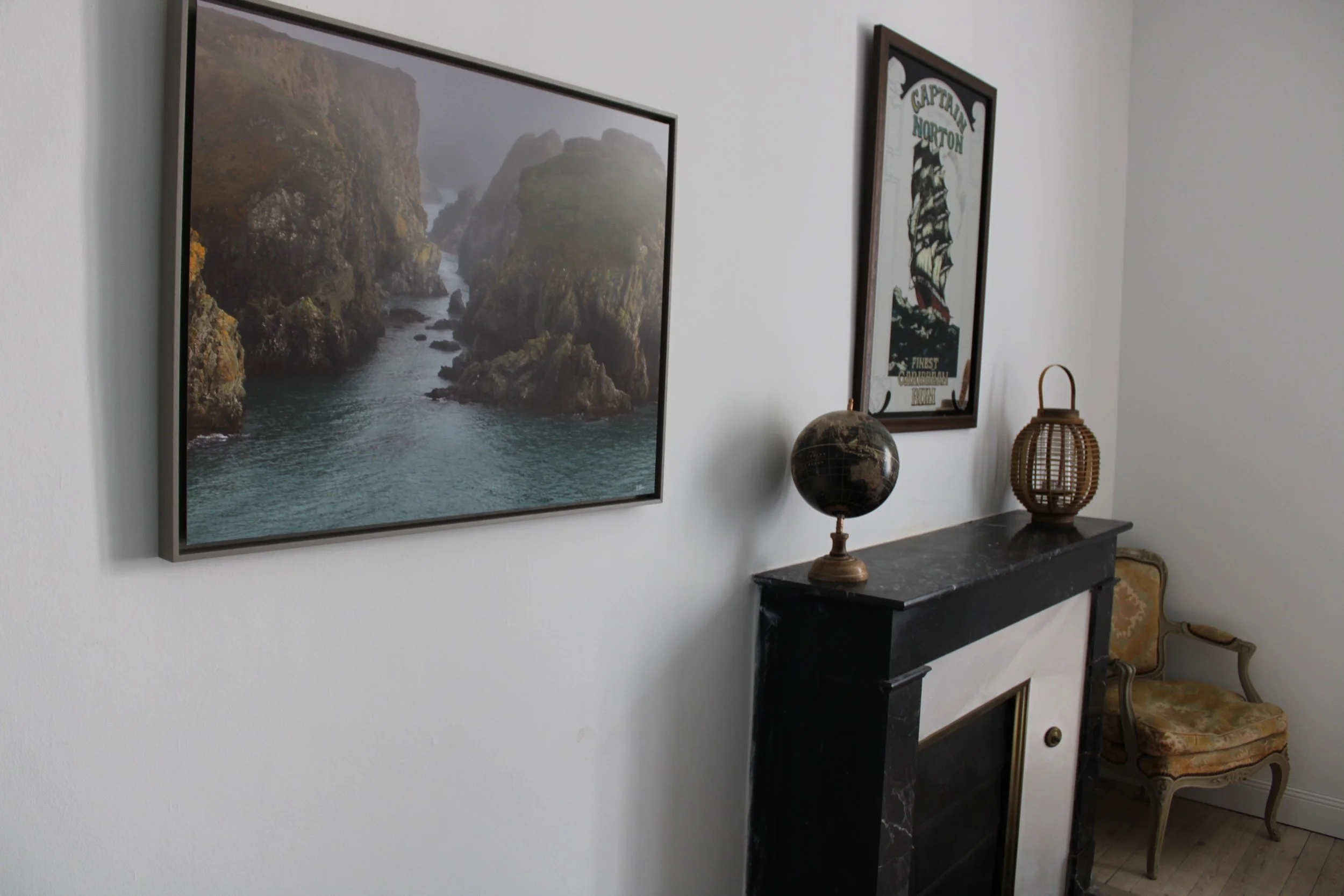 Une décoration intérieure avec deux tableaux accrochés au mur, une cheminée en marbre noir, une planète en globe terrestre, une lanterne en rotin, et un fauteuil ancien en bois avec un coussin.