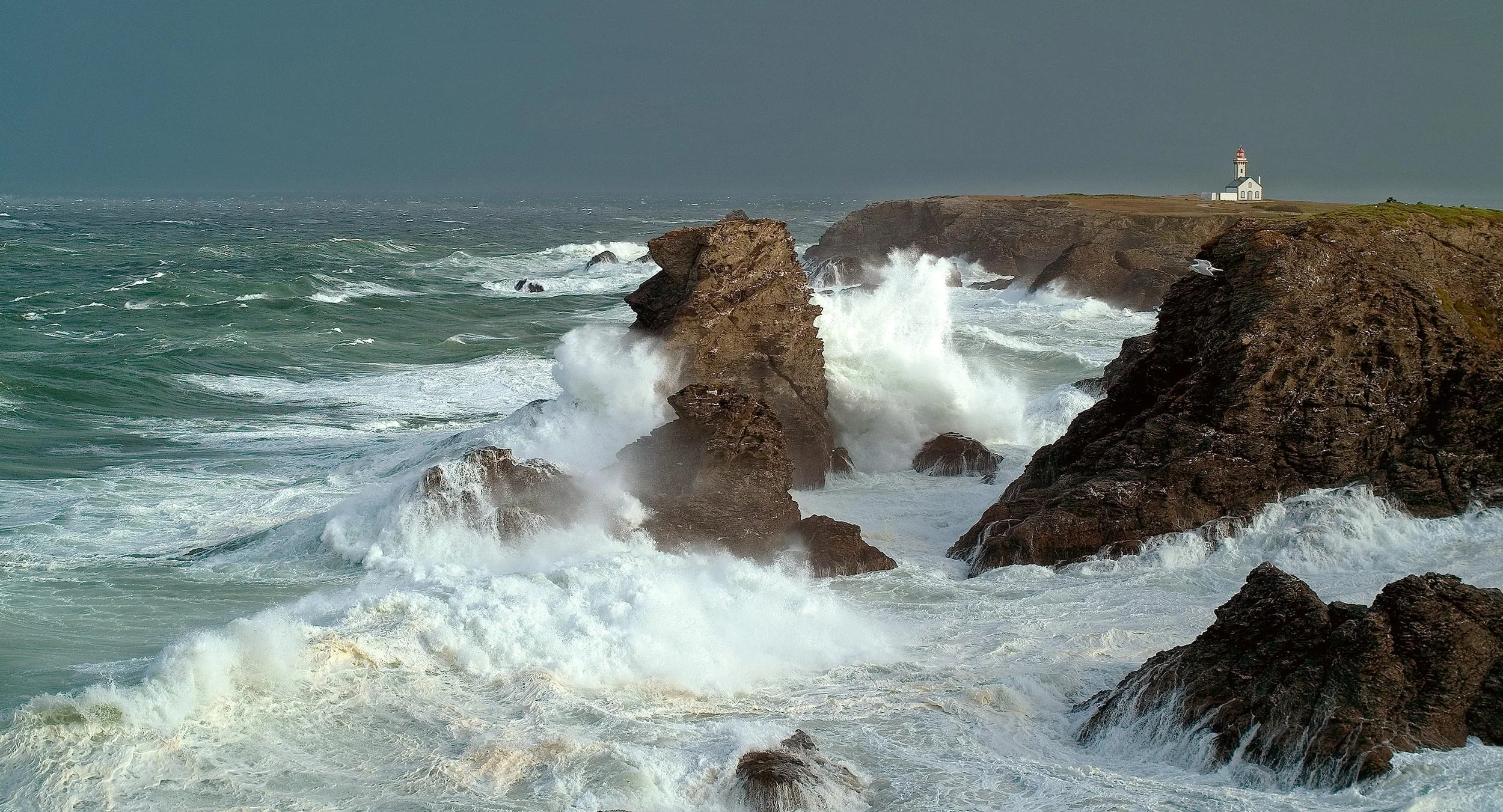 Un phare blanc situé sur une falaise au bord de la mer agitée avec des vagues s'écrasant contre les rochers.