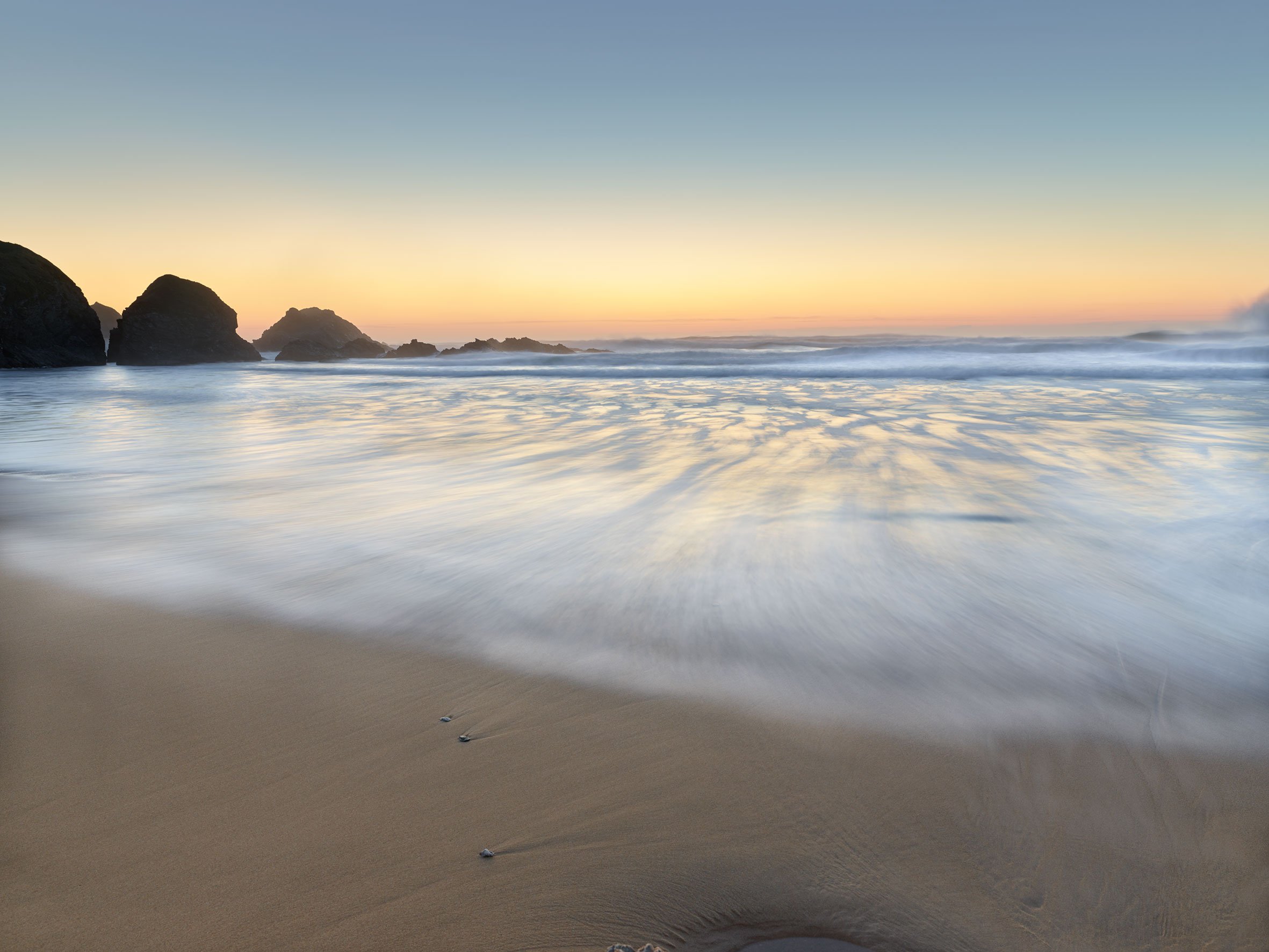 Plage de sable avec des rochers au loin et des vagues qui s'écrasent à l'horizon, lors d'un coucher ou lever de soleil.