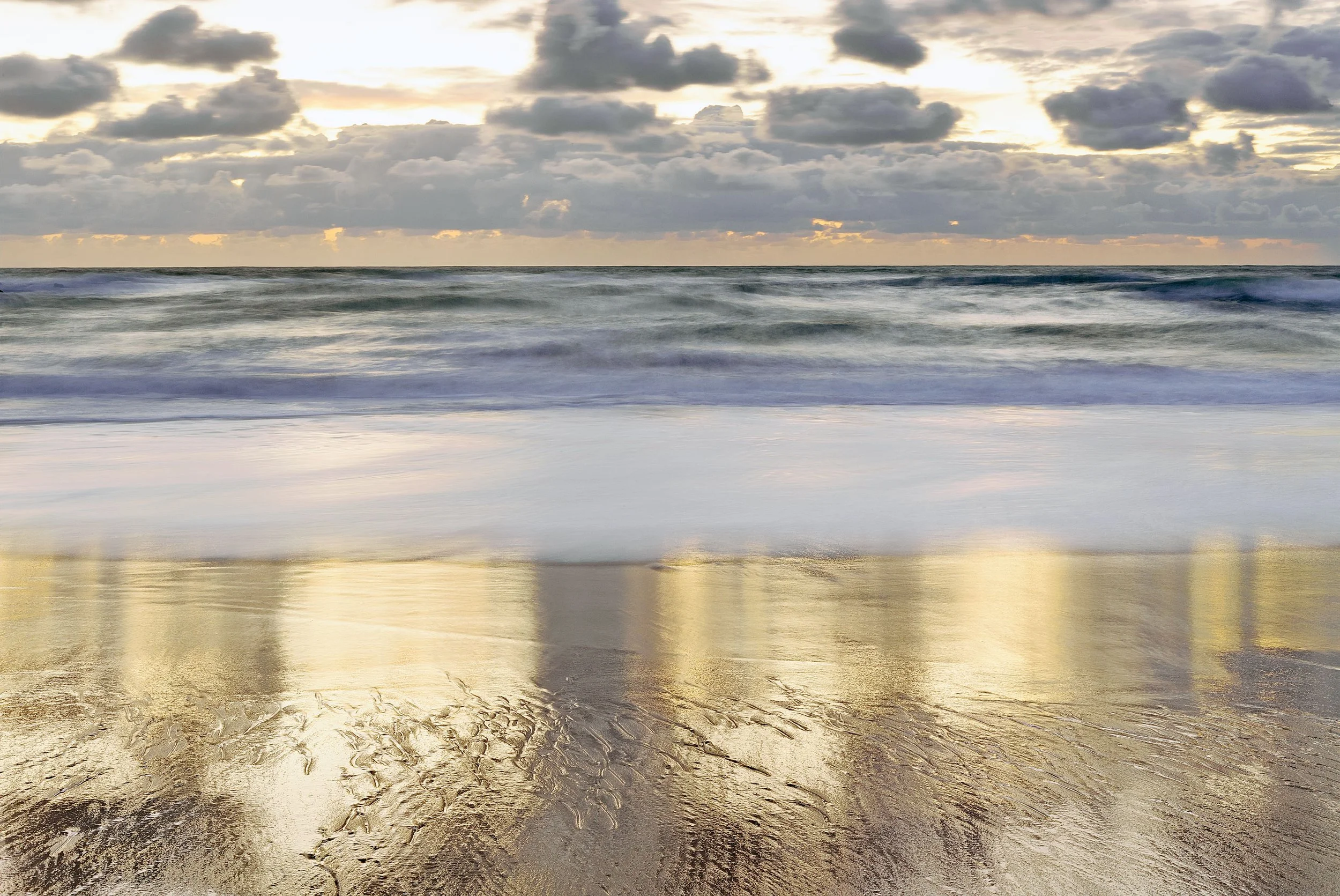 Vue d'une plage avec des vagues, un ciel nuageux et le reflet du coucher de soleil sur le sable humide.