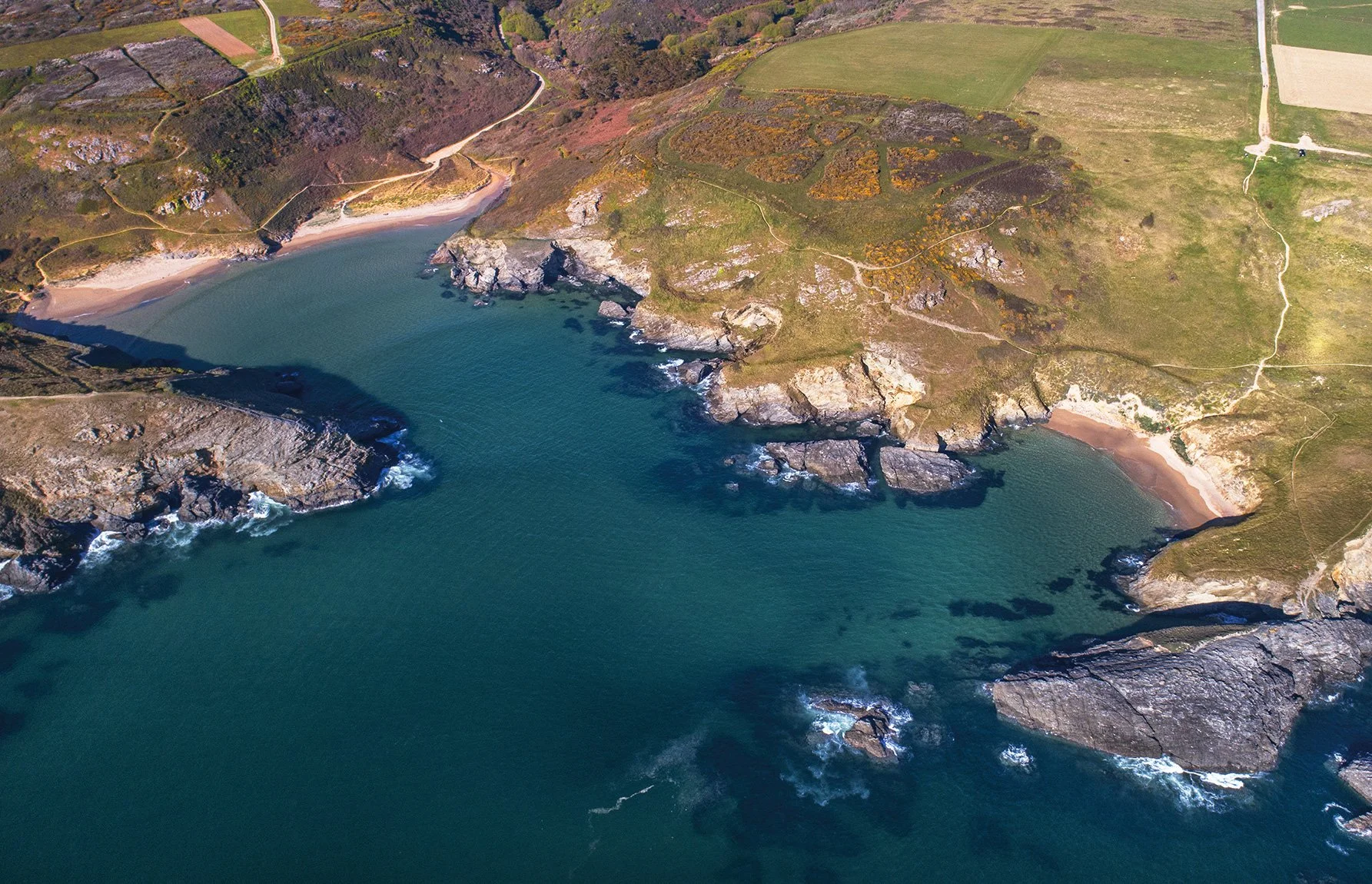 Vue aérienne de la côte avec des falaises rocheuses, des plages de sable et une mer turquoise entourée de collines verdoyantes.