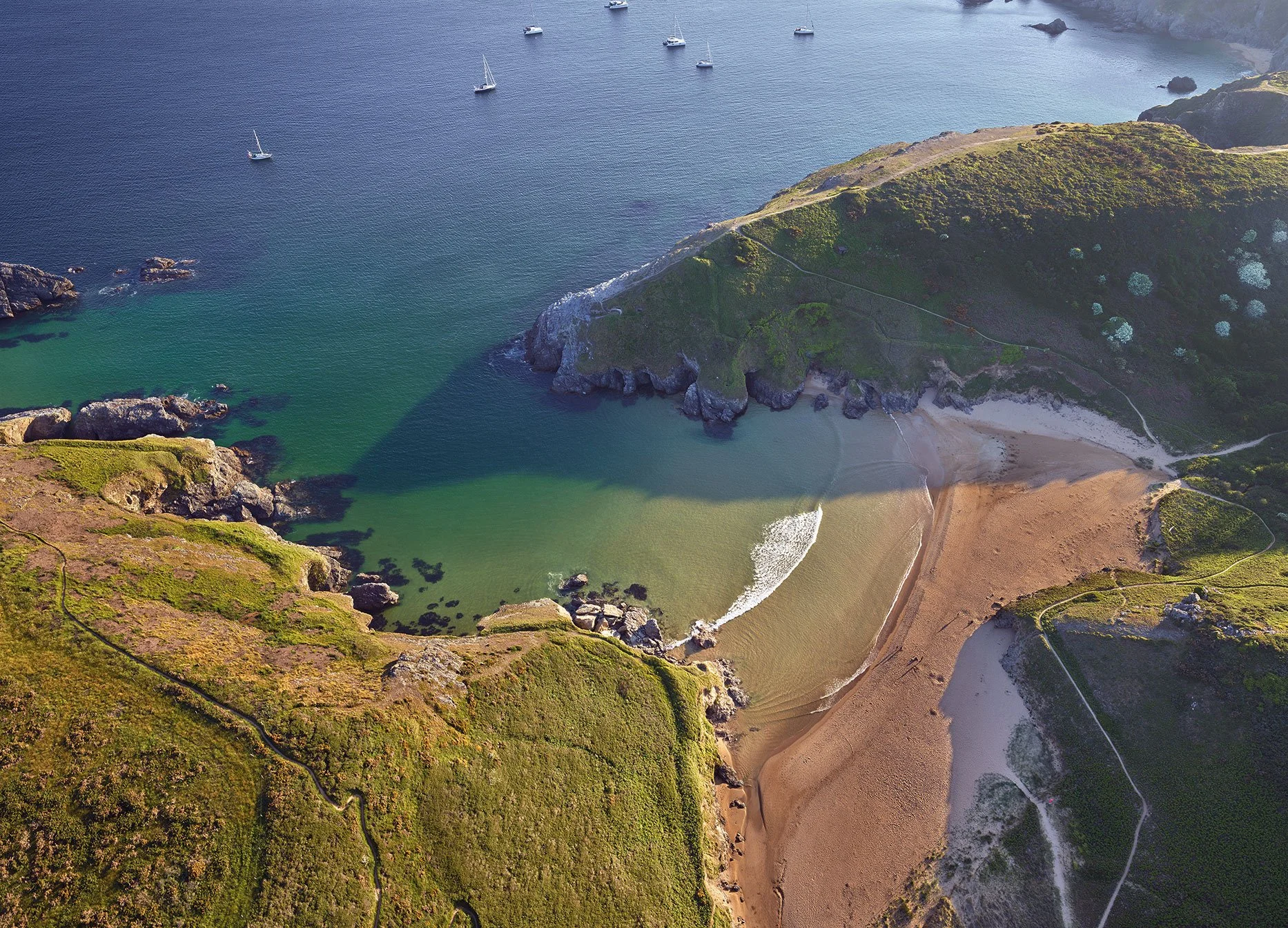 Vue aérienne d'une baie avec des eaux bleues et vertes, une plage de sable, des falaises recouvertes de végétation et plusieurs voiliers dans la mer.