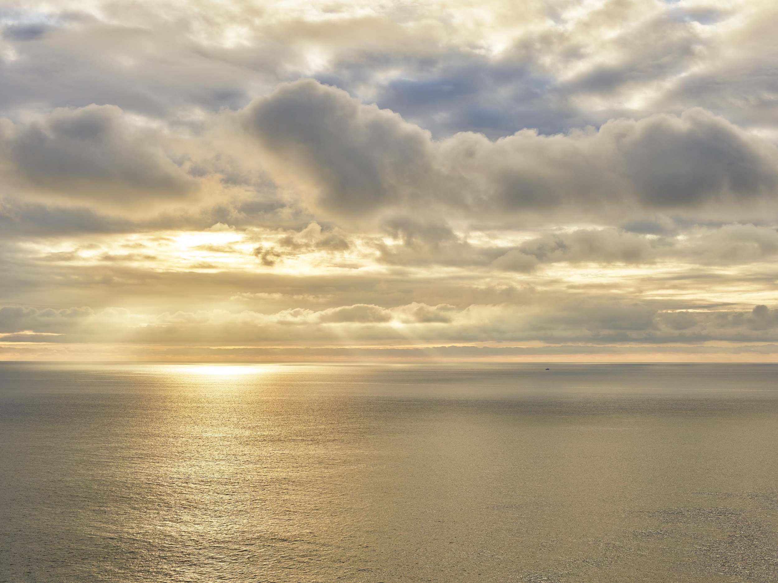 Ciel nuageux au-dessus de la mer lors du coucher ou lever du soleil, avec des nuages gris et une lumière dorée sur l'eau.