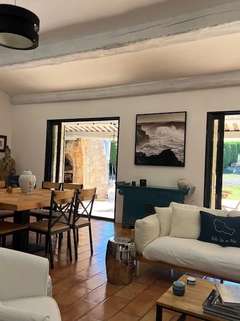 Salon lumineux avec un canapé blanc, une table basse, un tableau noir et blanc, une cheminée bleue, et des fenêtres donnant sur l'extérieur.
