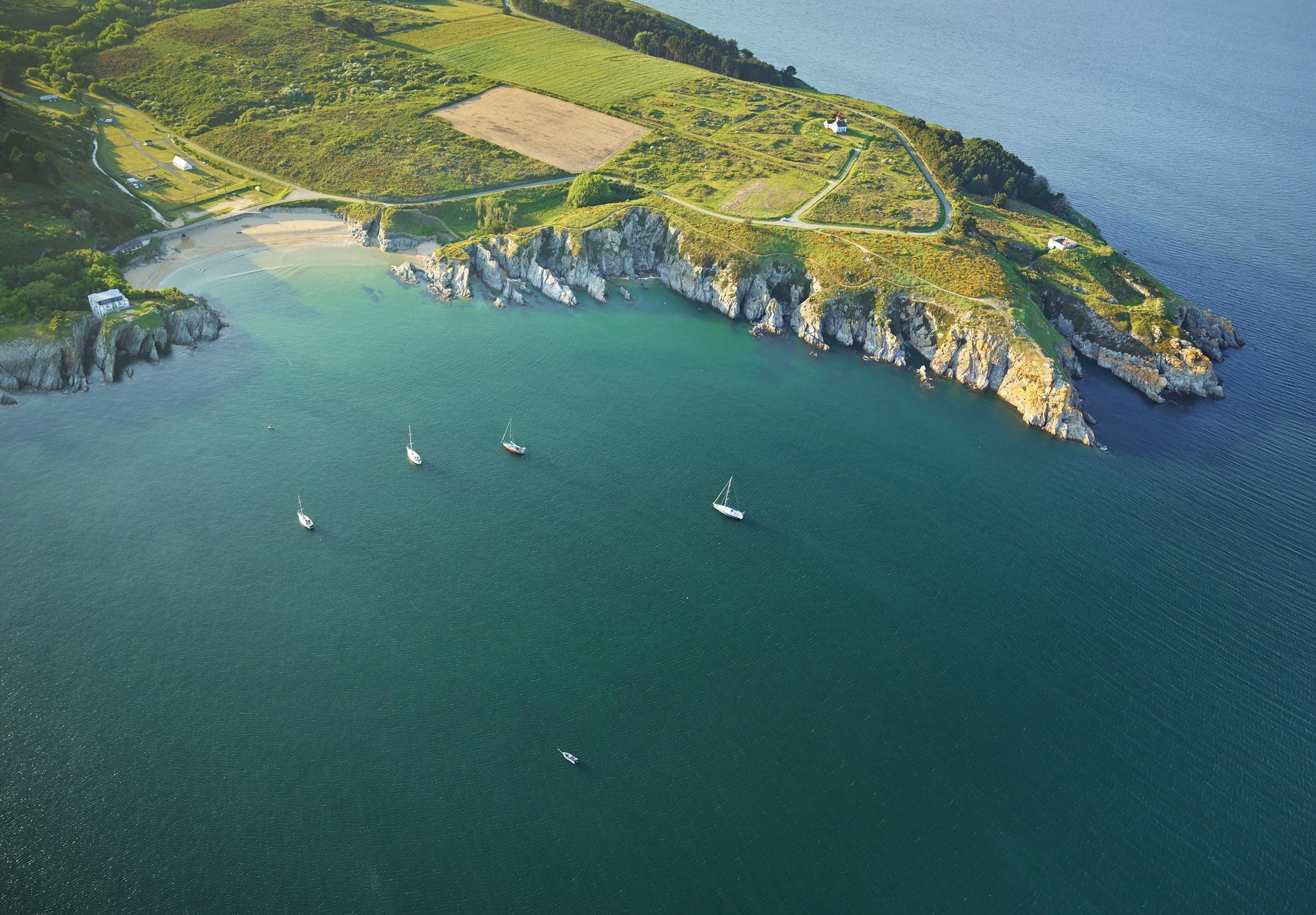 Vue aérienne d'une côte rocheuse entourée d'eau avec quelques bateaux en mer. Sur la côte, des herbes vertes, des champs, et quelques bâtiments, y compris une église avec un toit rouge, un petit chemin serpentant à travers la zone.