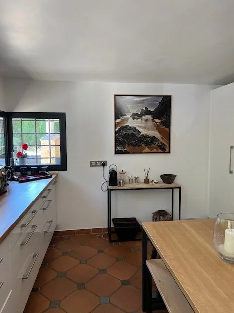 Cuisine avec un plan de travail en bois, une fenêtre avec des volets noirs, une peinture de paysage côtier accroché au mur, et une petite table avec des objets décoratifs.