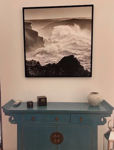 Une pièce avec un tableau noir et blanc d'une scène de vagues s'écrasant contre des rochers, au-dessus d'une console bleue avec des objets décoratifs, dont un vase et une boîte.