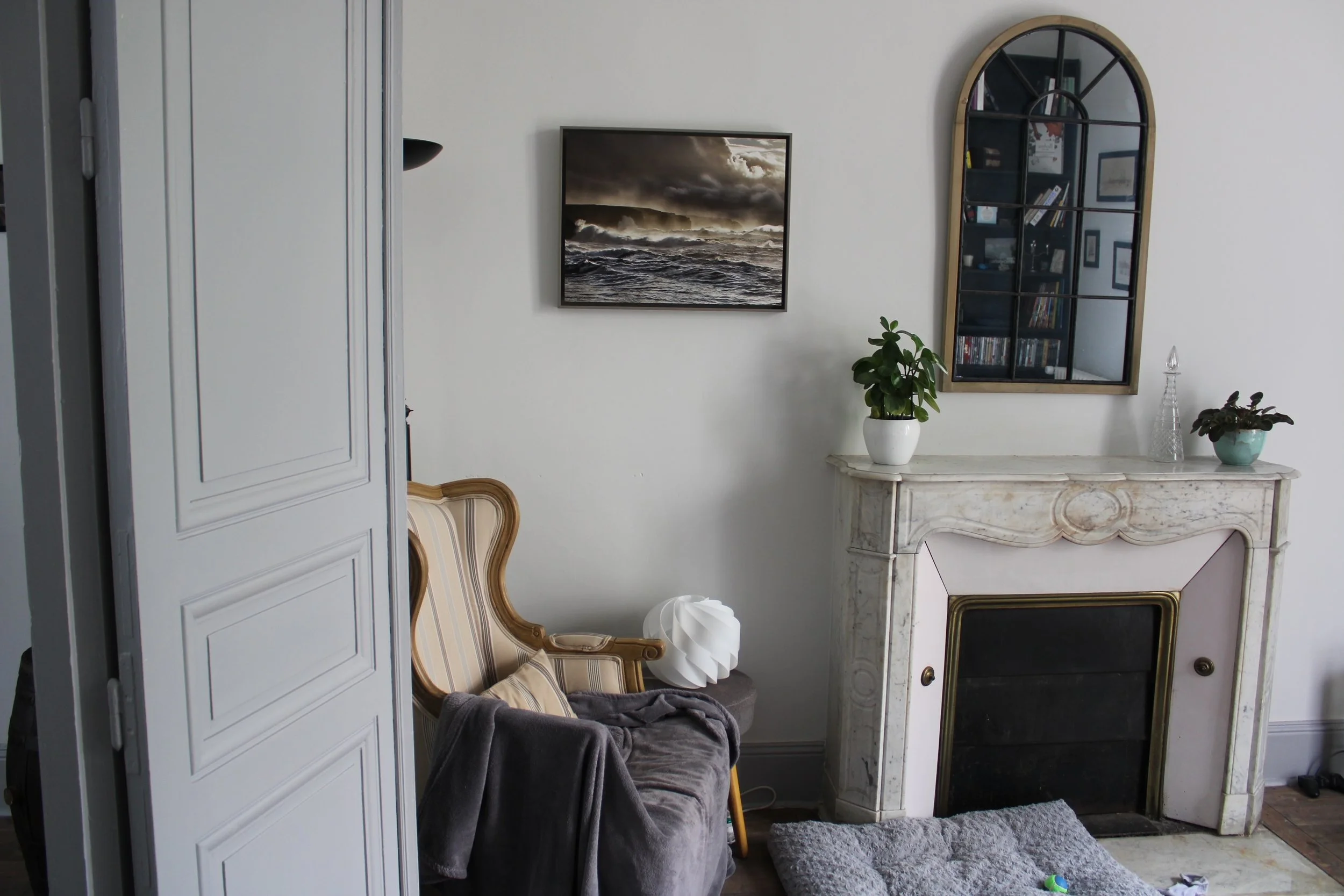 Intérieur d'un salon avec un fauteuil, un manteau sur le fauteuil, un miroir en arc au mur, une plante en pot, une cheminée en marbre, une peinture représentant une mer tempétueuse, et une étagère avec des livres derrière le miroir.