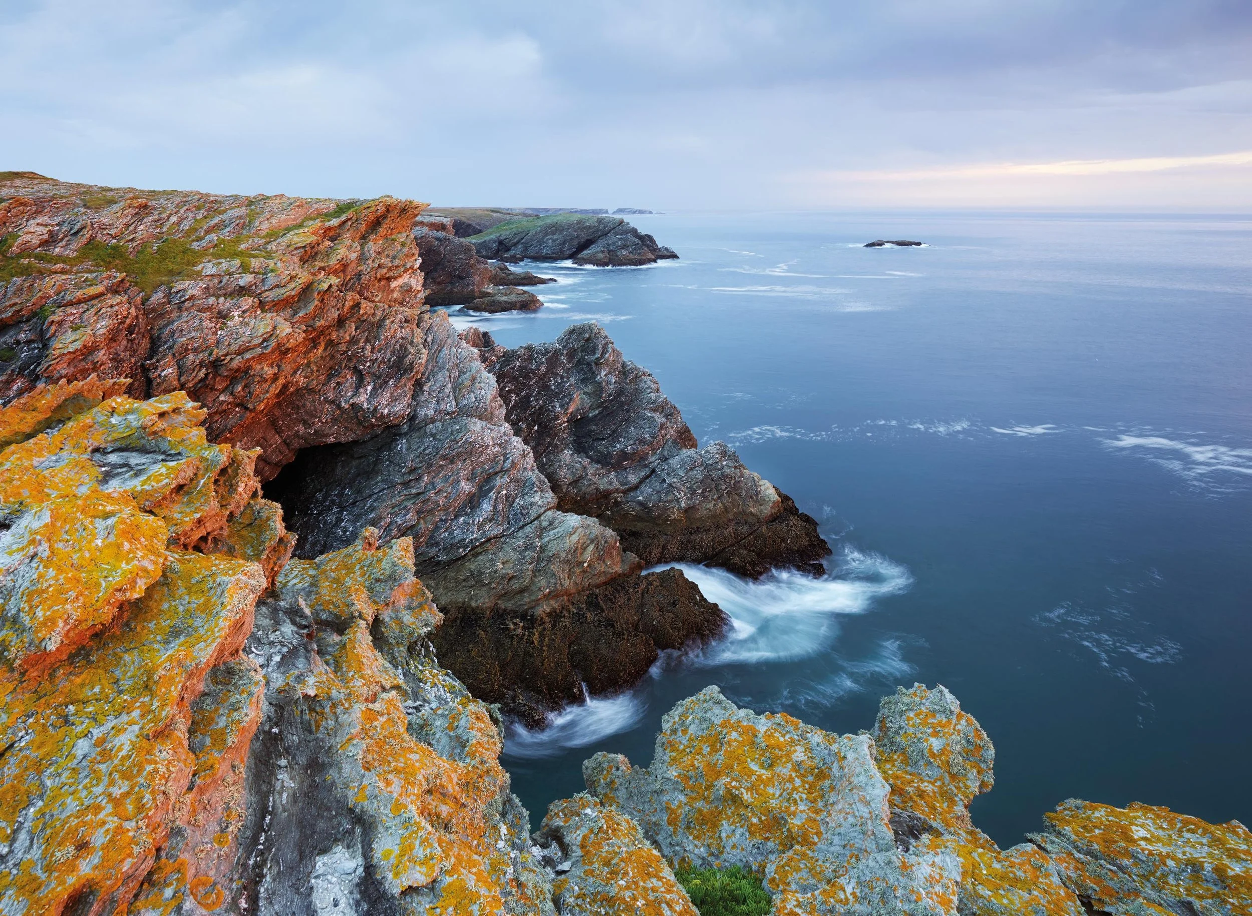 Falaises rocheuses colorées en premier plan avec l'océan au fond et un ciel nuageux.
