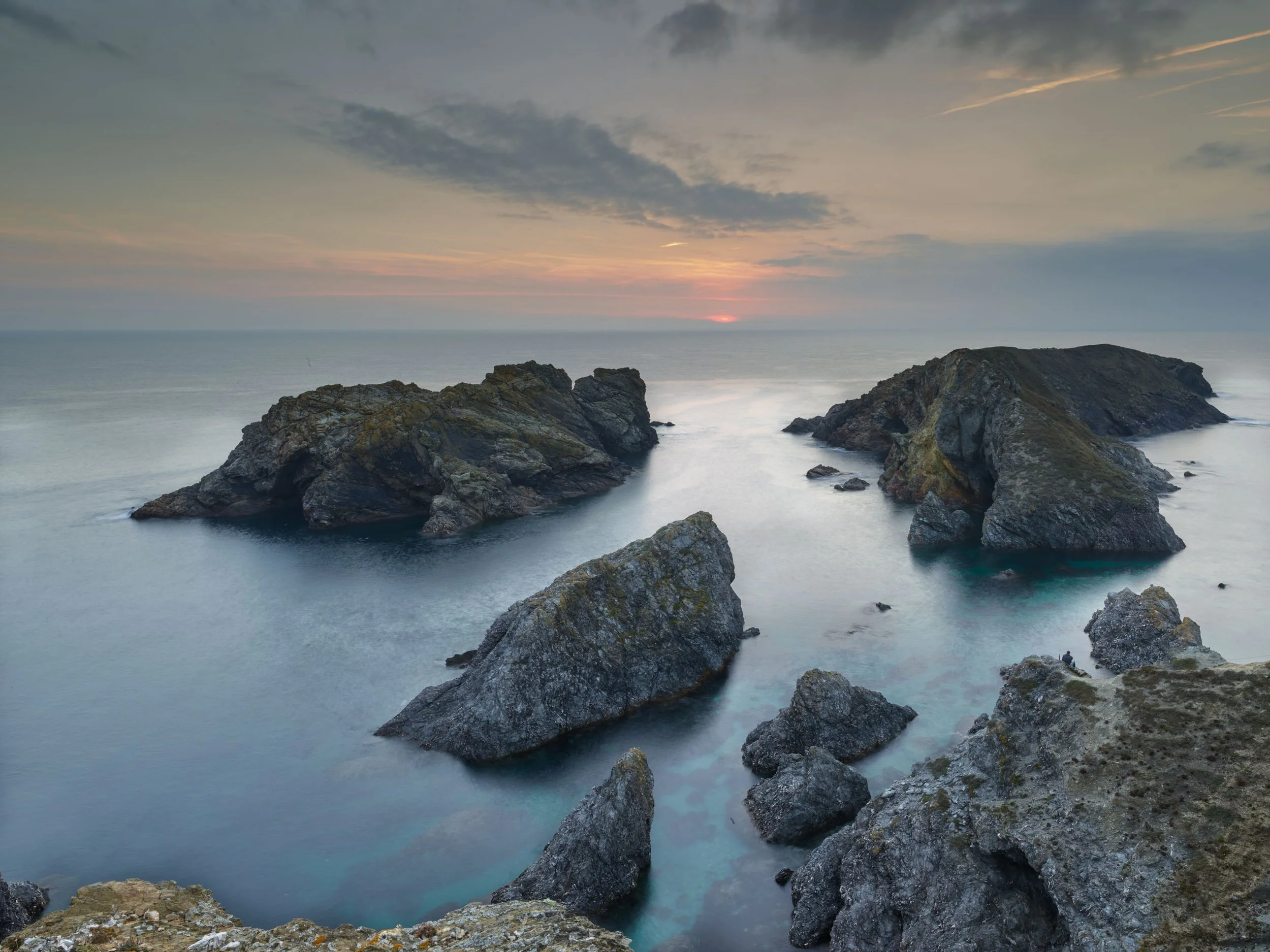 Coucher du soleil sur des rochers au bord de la mer, avec un ciel partiellement nuageux.