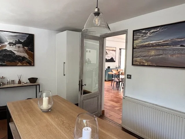 Intérieur d'une salle à manger avec table en bois, deux bougies dans des verres, tableaux muraux représentant des plages, porte en verre menant à une autre pièce avec cheminée.